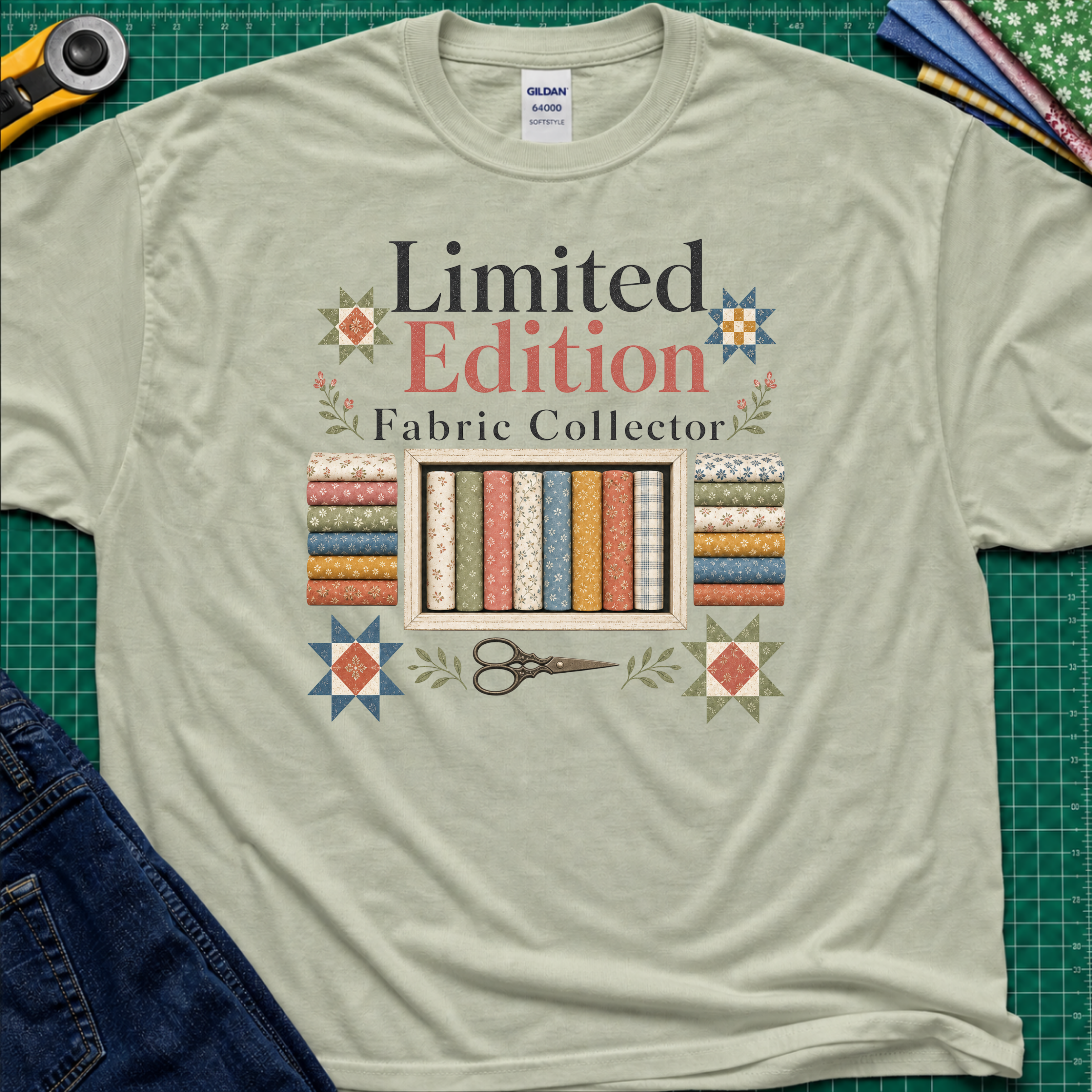 A light sage green Gildan t-shirt featuring a colorful graphic that reads 'Limited Edition Fabric Collector' with fabric rolls, scissors, and decorative stars. The shirt is laying on a green cutting mat with quilting supplies and blue jeans around the outside.