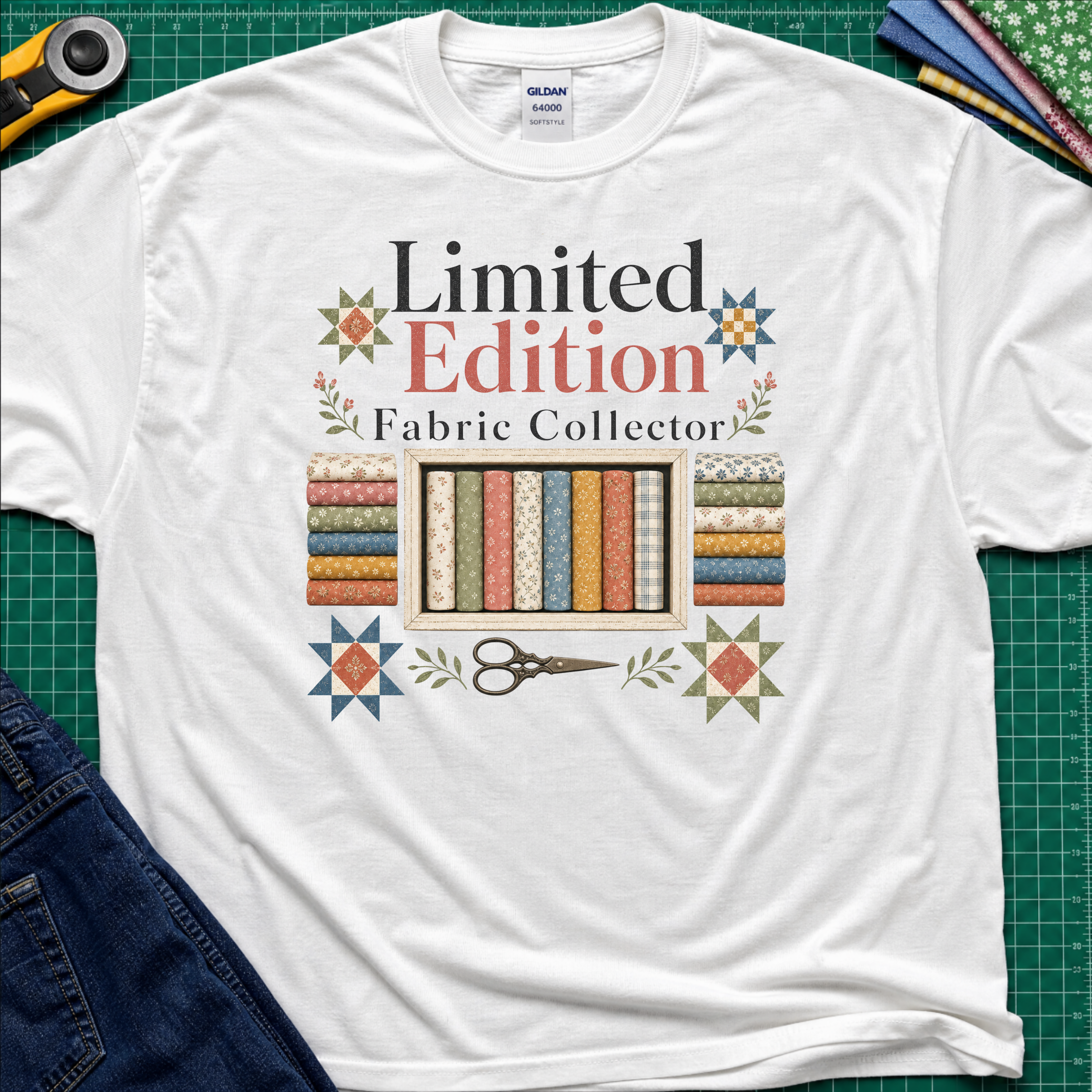 A white Gildan t-shirt featuring a colorful graphic that reads 'Limited Edition Fabric Collector' with fabric rolls, scissors, and decorative stars. The shirt is laying on a green cutting mat with quilting supplies and blue jeans around the outside.