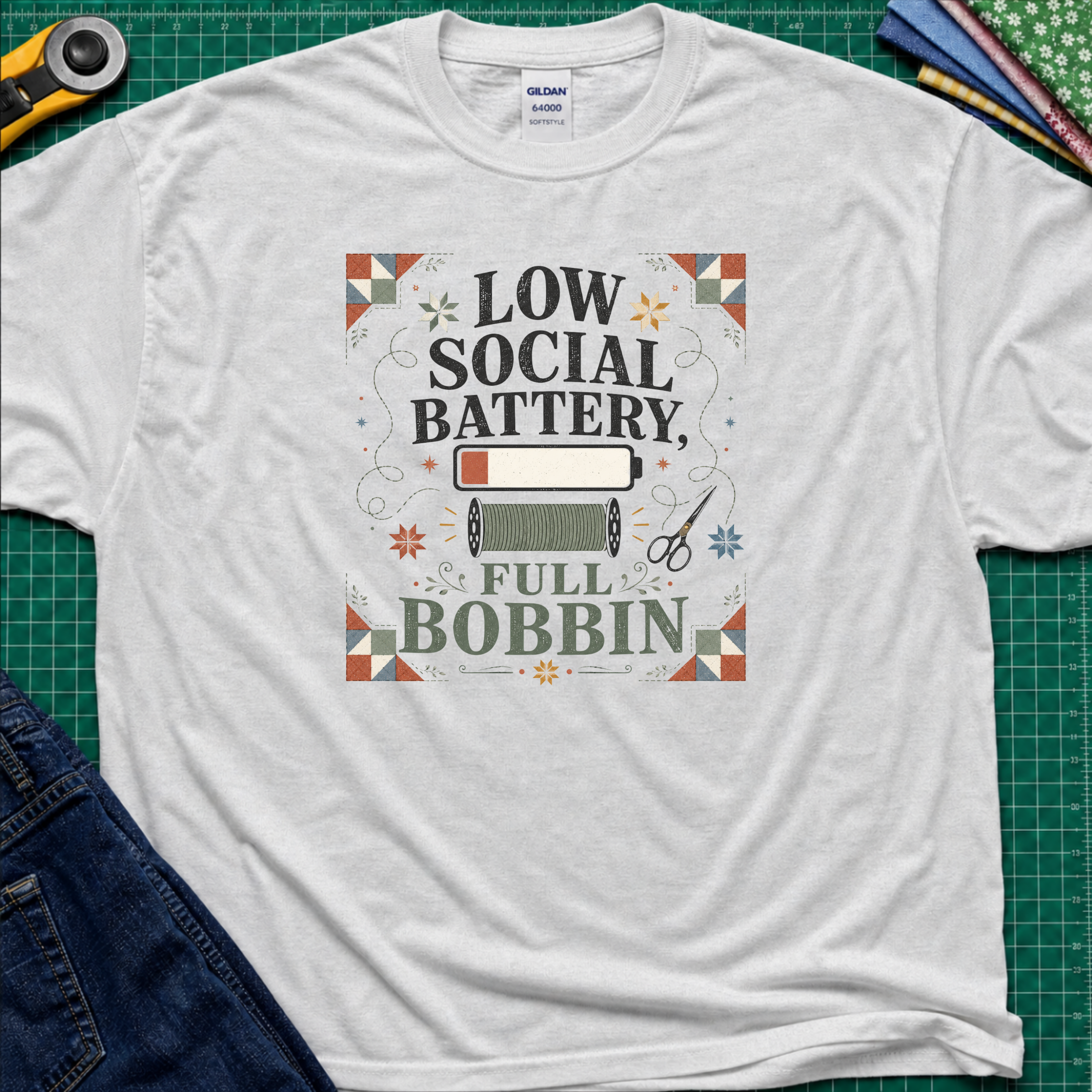 A light grey Gildan t-shirt with a retro-style graphic that reads 'LOW SOCIAL BATTERY, FULL BOBBIN' surrounded by a quilt-like border and sewing motifs, presented in a flat-lay on a green cutting mat with crafting props.