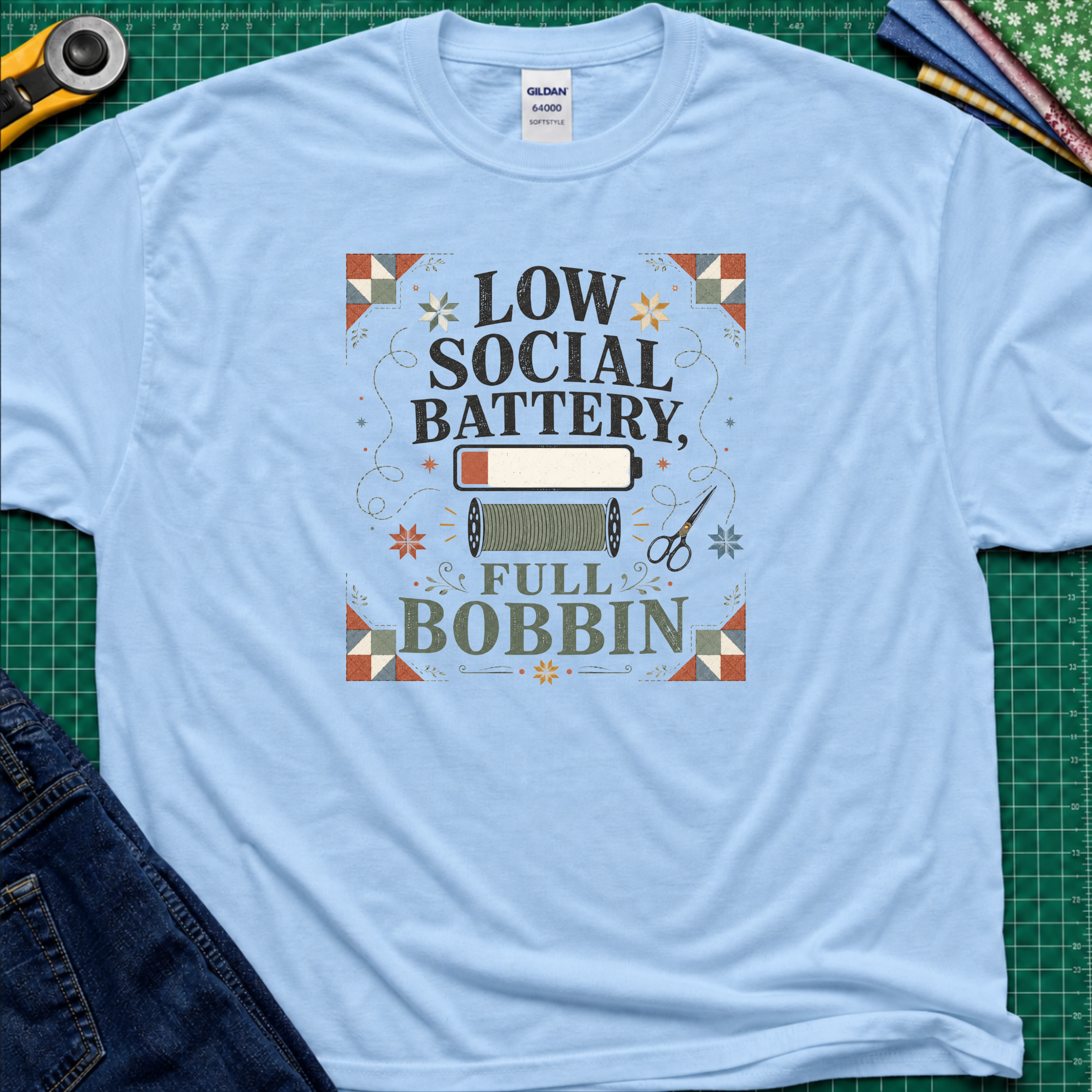 A light blue Gildan t-shirt with a retro-style graphic that reads 'LOW SOCIAL BATTERY, FULL BOBBIN' surrounded by a quilt-like border and sewing motifs, presented in a flat-lay on a green cutting mat with crafting props.