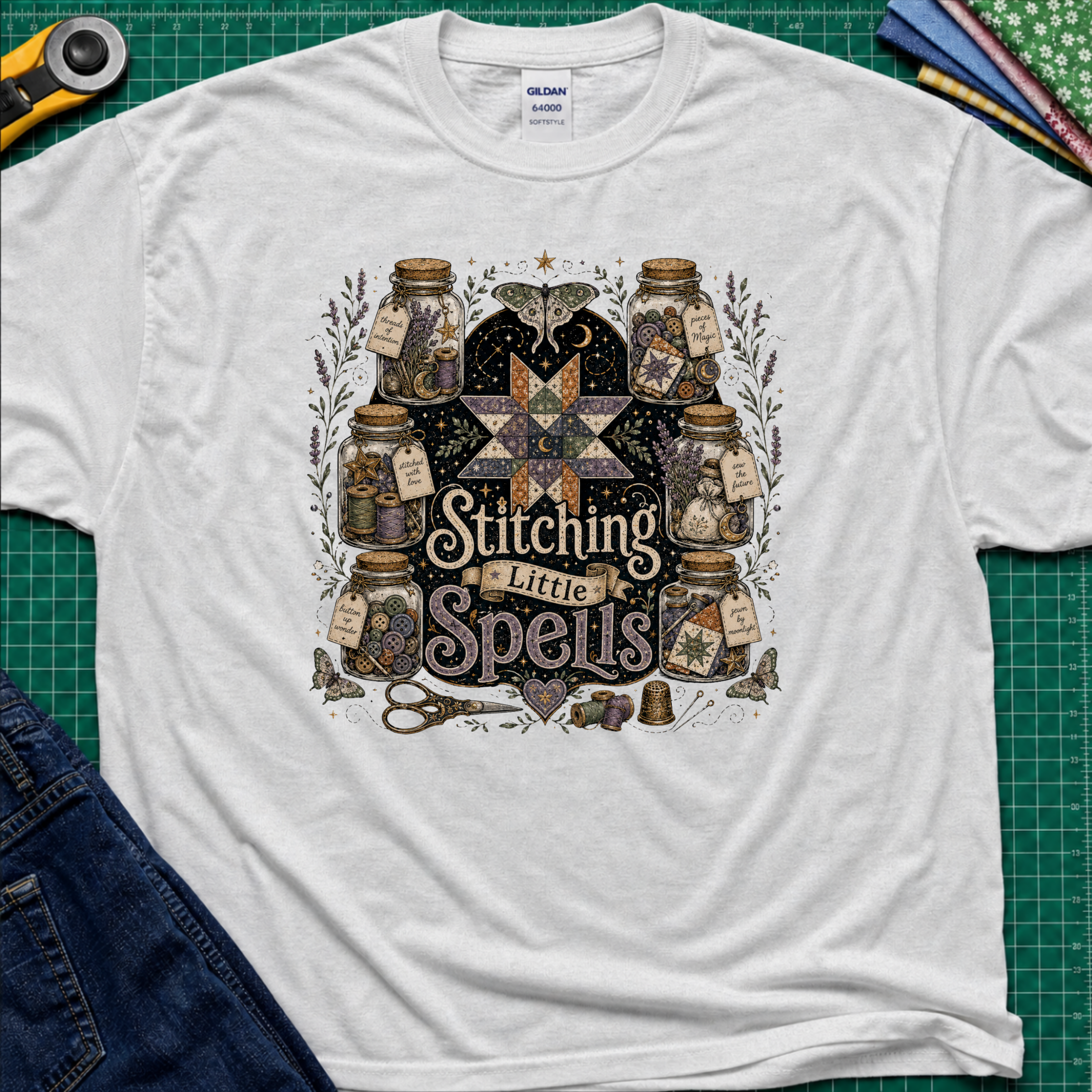 A light grey Gildan t-shirt laid flat on a green cutting mat, featuring a detailed graphic of vintage glass jars, quills, scissors, and a star with the text 'Stitching Little Spells'.