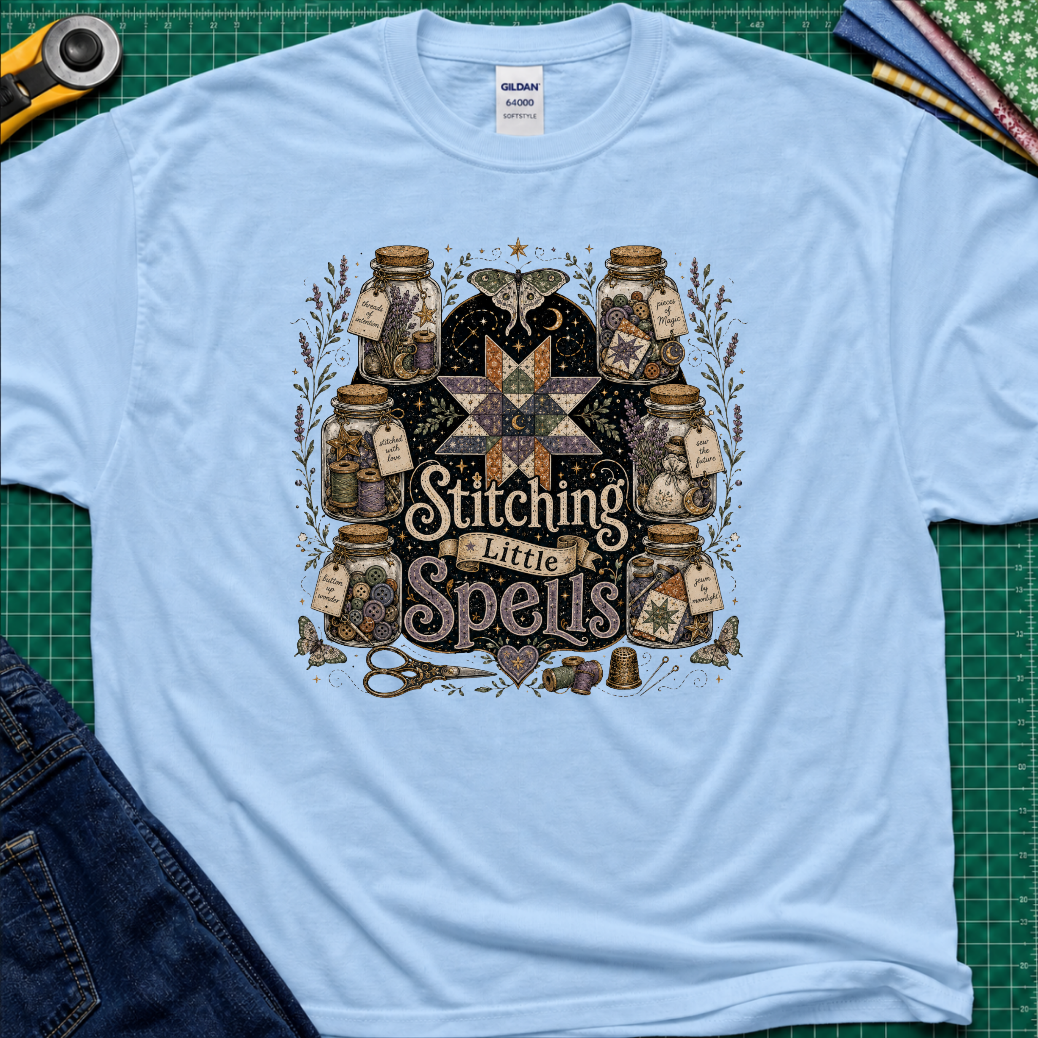 A light blue Gildan t-shirt laid flat on a green cutting mat, featuring a detailed graphic of vintage glass jars, quills, scissors, and a star with the text 'Stitching Little Spells'.