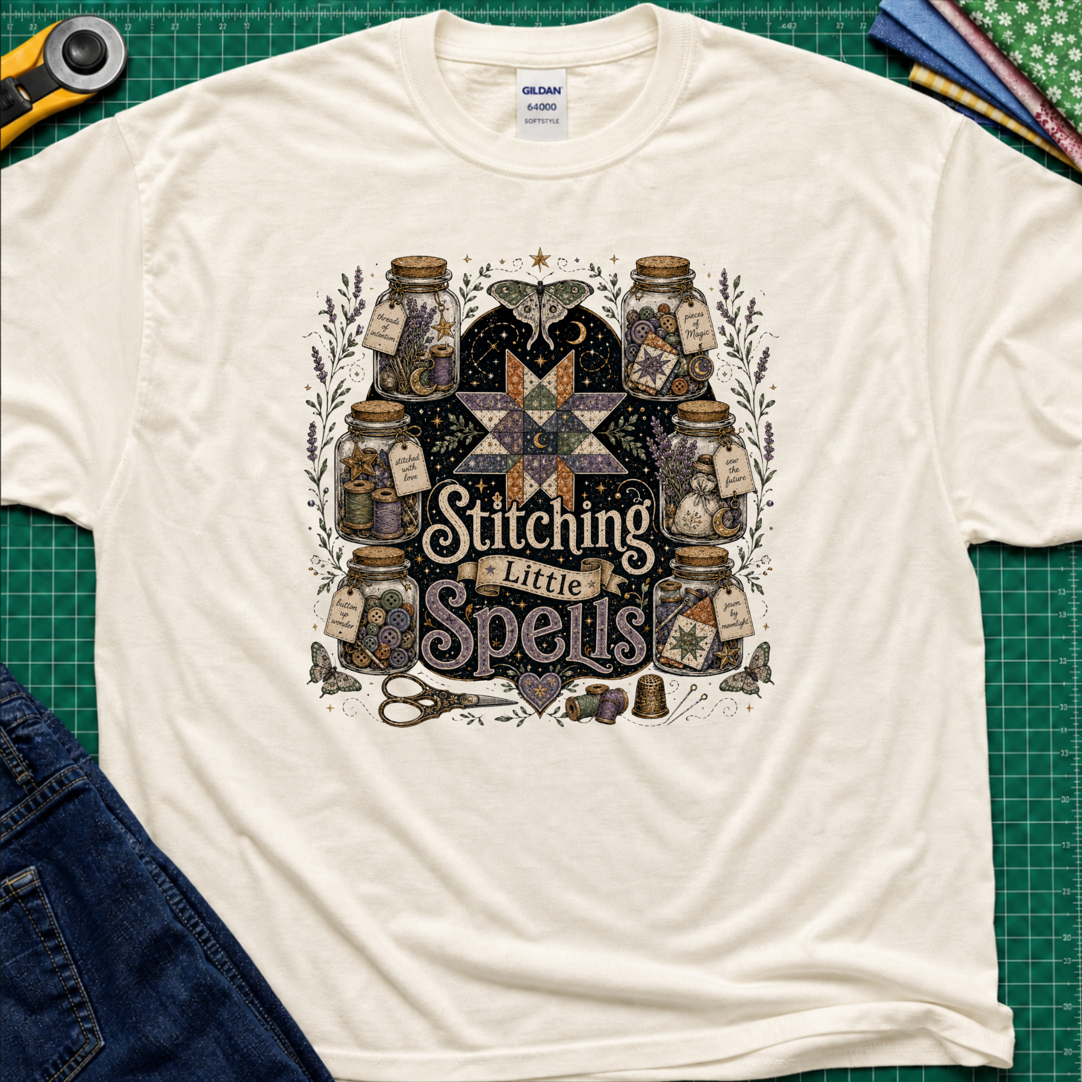 A light cream Gildan t-shirt laid flat on a green cutting mat, featuring a detailed graphic of vintage glass jars, quills, scissors, and a star with the text 'Stitching Little Spells'.