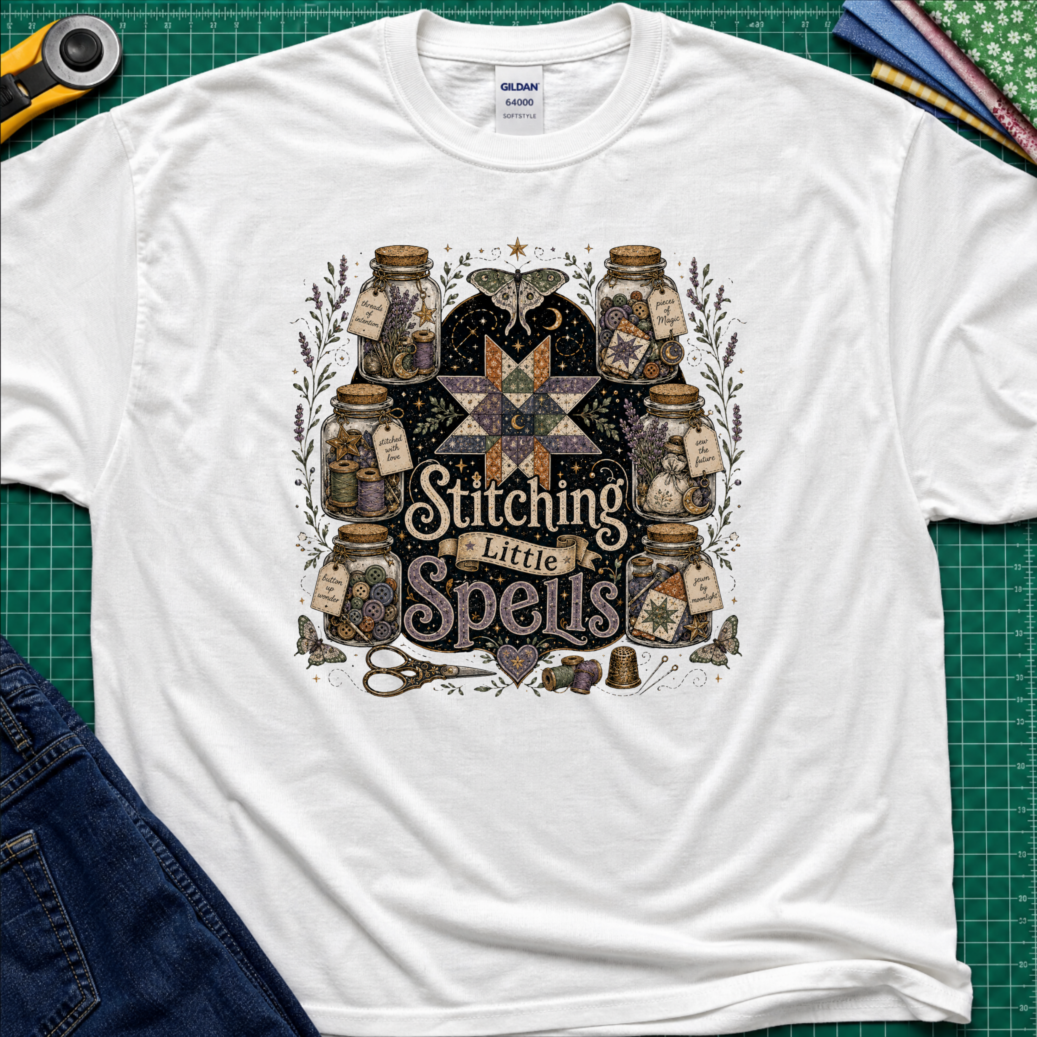 A white Gildan t-shirt laid flat on a green cutting mat, featuring a detailed graphic of vintage glass jars, quills, scissors, and a star with the text 'Stitching Little Spells'.
