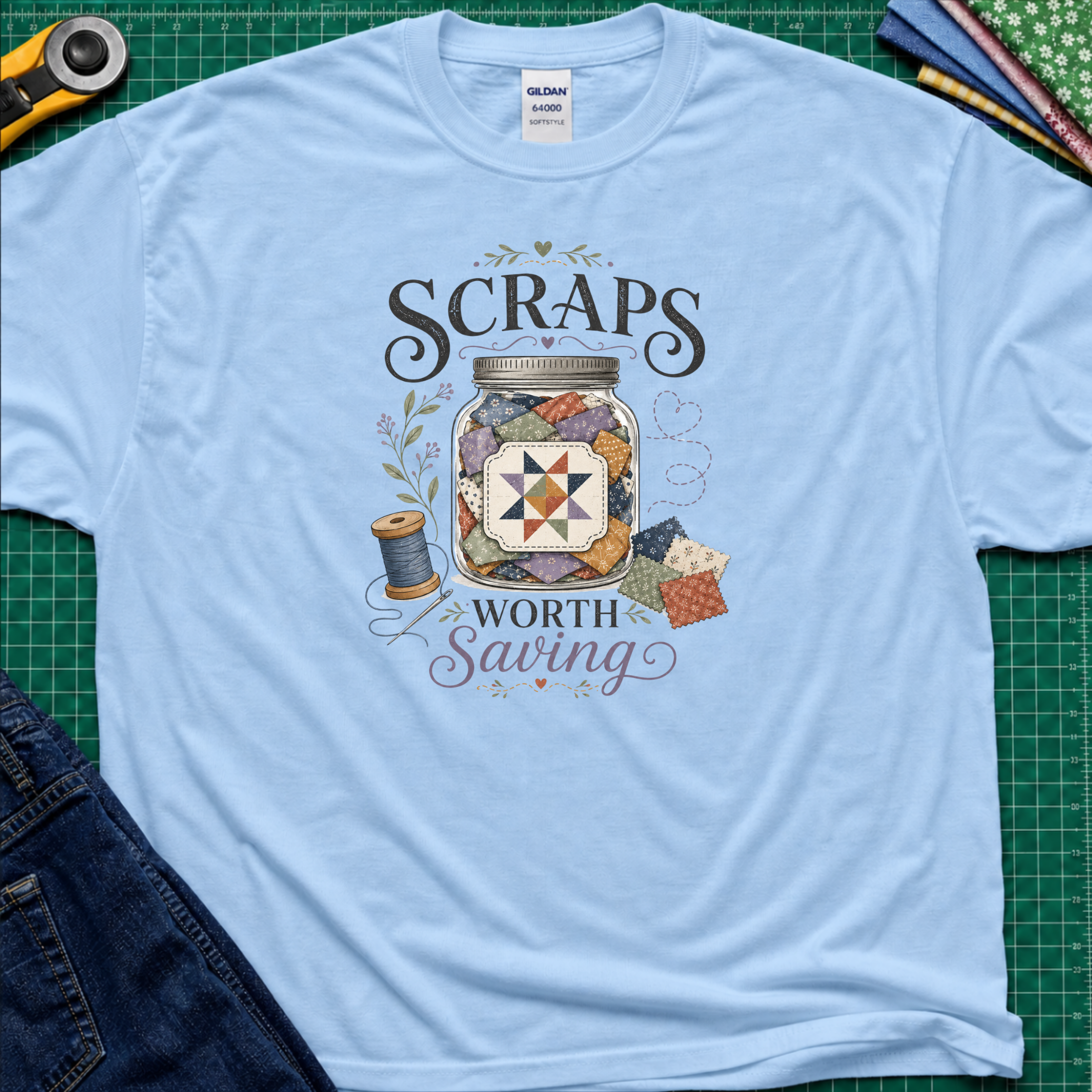A light blue Gildan t-shirt laid flat on a green cutting mat, featuring a graphic design that reads 'SCRAPS WORTH Saving' around an illustration of a jar filled with scraps, thread spool, and fabric pieces. A rotary cutter, scissors, and folded fabrics are visible around the shirt as styling props.