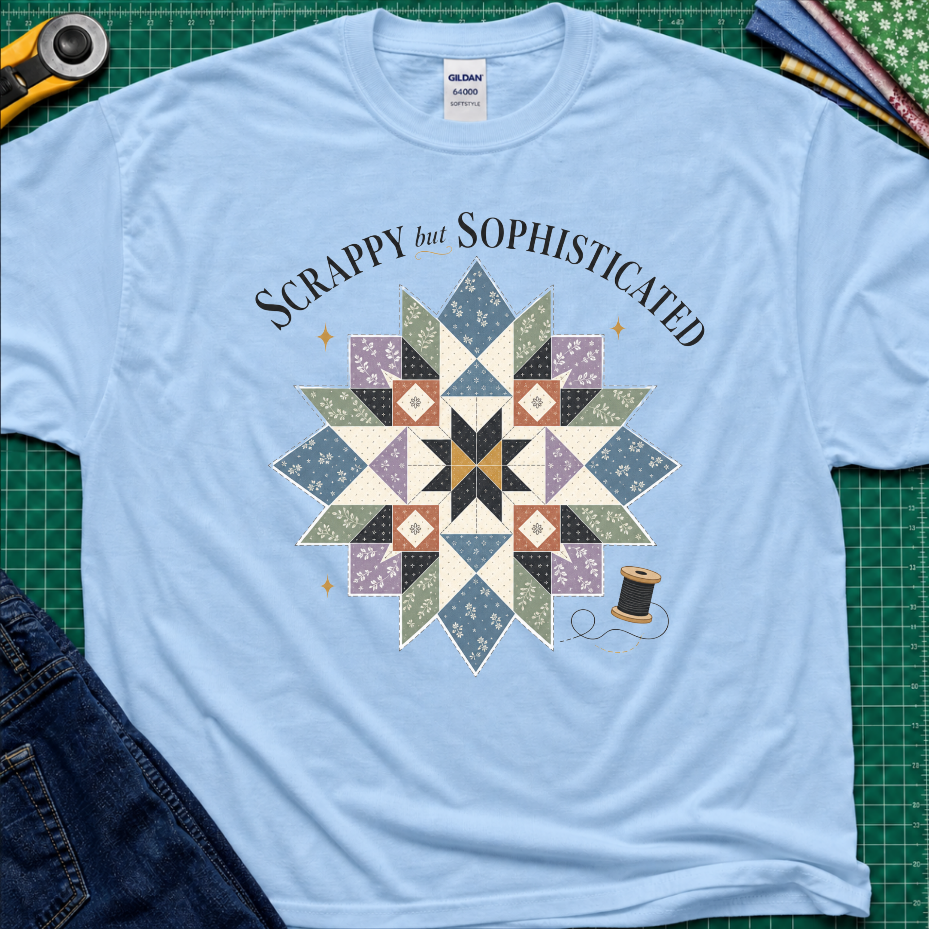 A light blue Gildan t-shirt with a colorful quilted starburst graphic and the text 'Scrappy but Sophisticated' printed on the front, styled on a green cutting mat with sewing tools and denim.