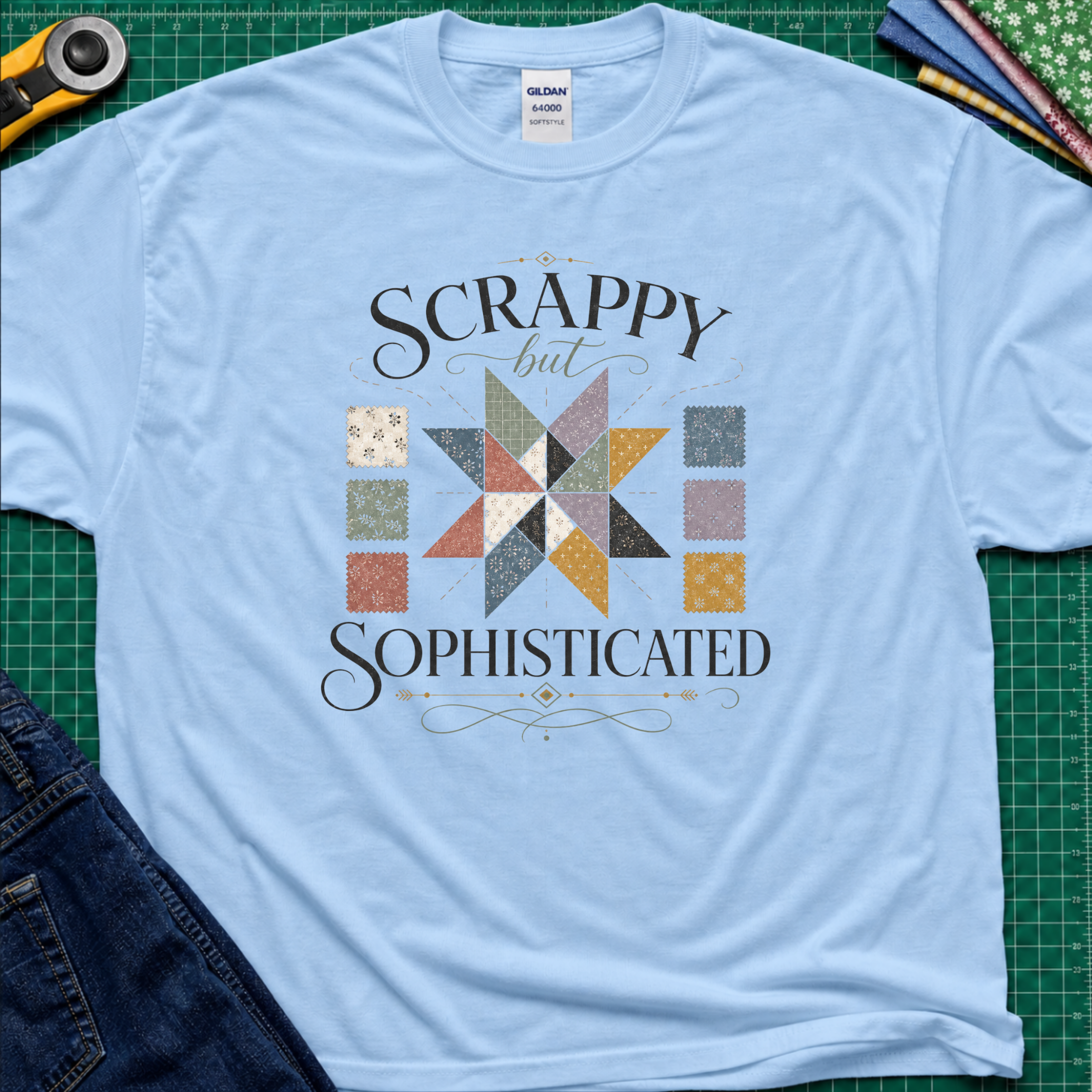 Alight blue Gildan t-shirt laid flat on a green cutting mat background, featuring a colorful quilted star graphic and the words 'SCRAPPY but SOPHISTICATED' in black script. A rotary cutter, fabric, and jeans are partially visible as styling props.