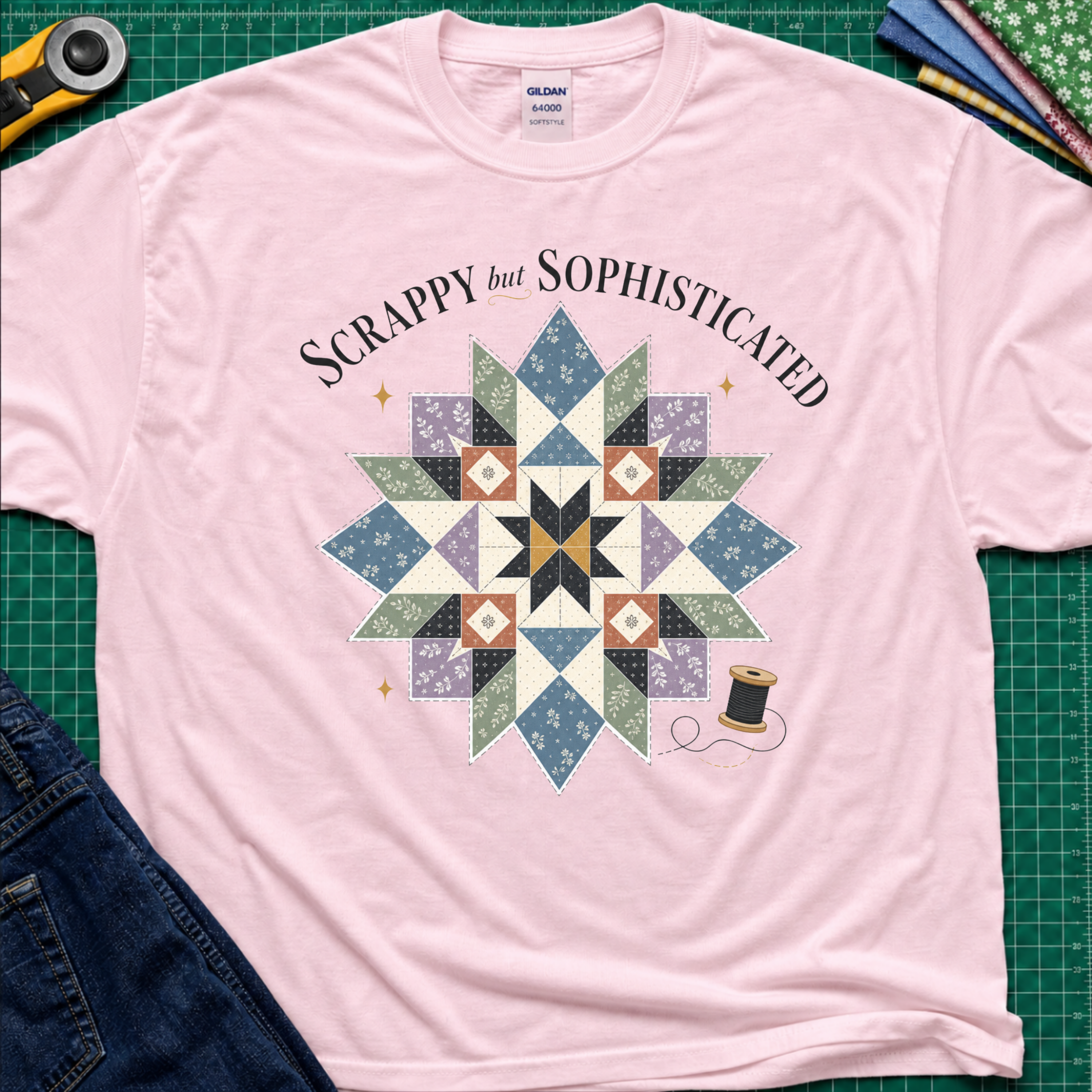 A light pink Gildan t-shirt with a colorful quilted starburst graphic and the text 'Scrappy but Sophisticated' printed on the front, styled on a green cutting mat with sewing tools and denim.