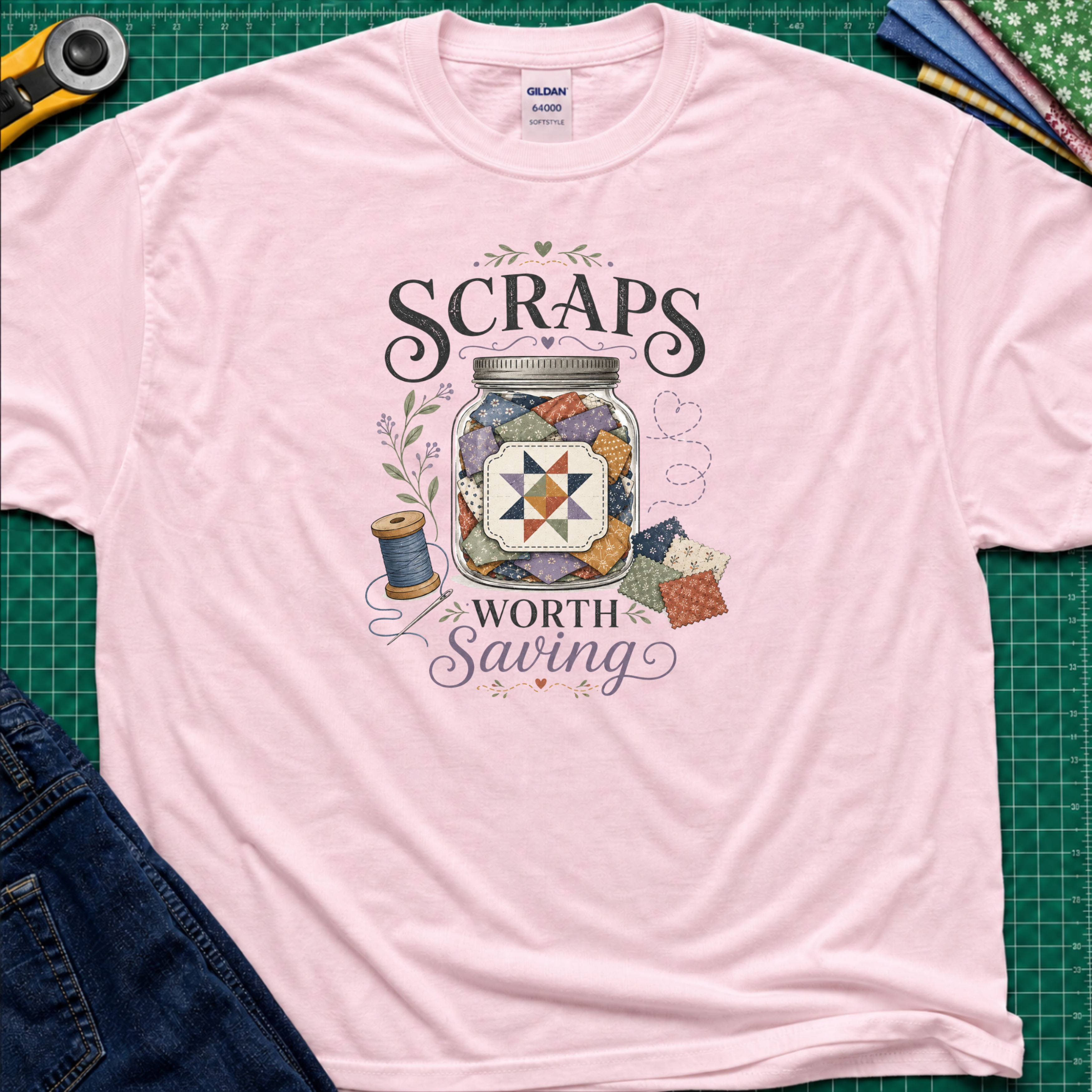 A light pink Gildan t-shirt laid flat on a green cutting mat, featuring a graphic design that reads 'SCRAPS WORTH Saving' around an illustration of a jar filled with scraps, thread spool, and fabric pieces. A rotary cutter, scissors, and folded fabrics are visible around the shirt as styling props.