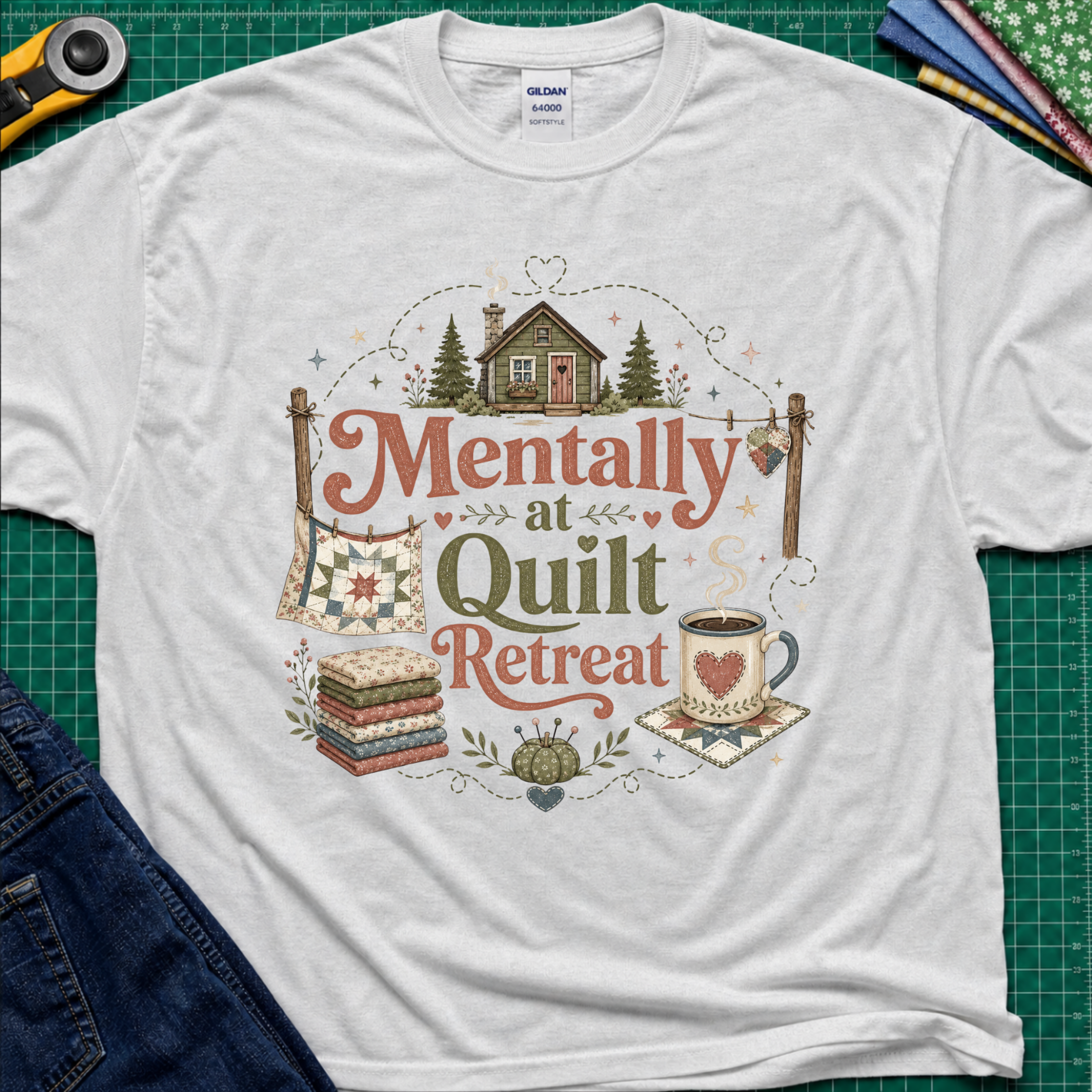 A light grey Gildan t-shirt with a rustic  style graphic reading 'Mentally at Quilt Retreat' surrounded by quilts, a cozy cabin, a coffee cup, and a stack of quilts. The shirt is styled flat on a green cutting mat with a rotary cutter, blue jeans and fabric stack.