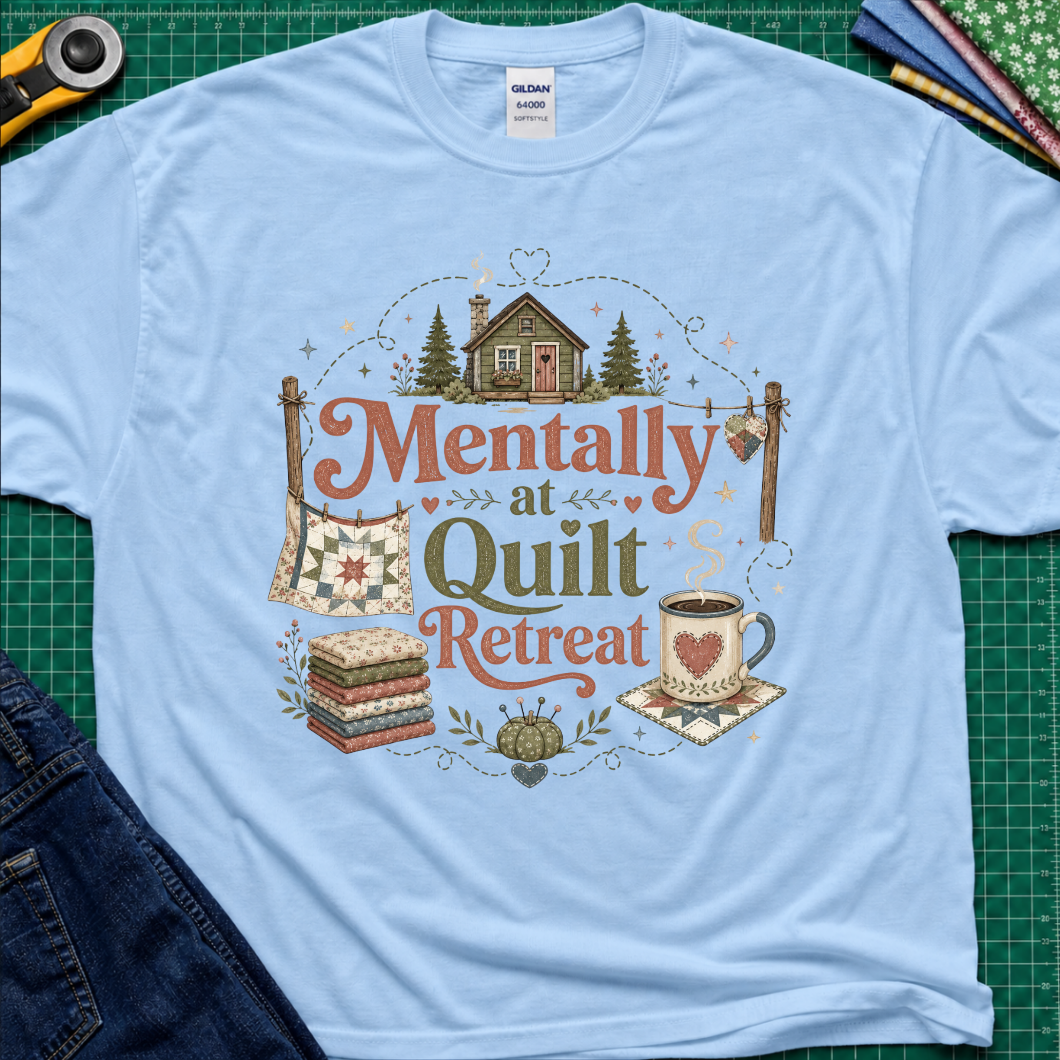 A light blue Gildan t-shirt with a rustic  style graphic reading 'Mentally at Quilt Retreat' surrounded by quilts, a cozy cabin, a coffee cup, and a stack of quilts. The shirt is styled flat on a green cutting mat with a rotary cutter, blue jeans and fabric stack.