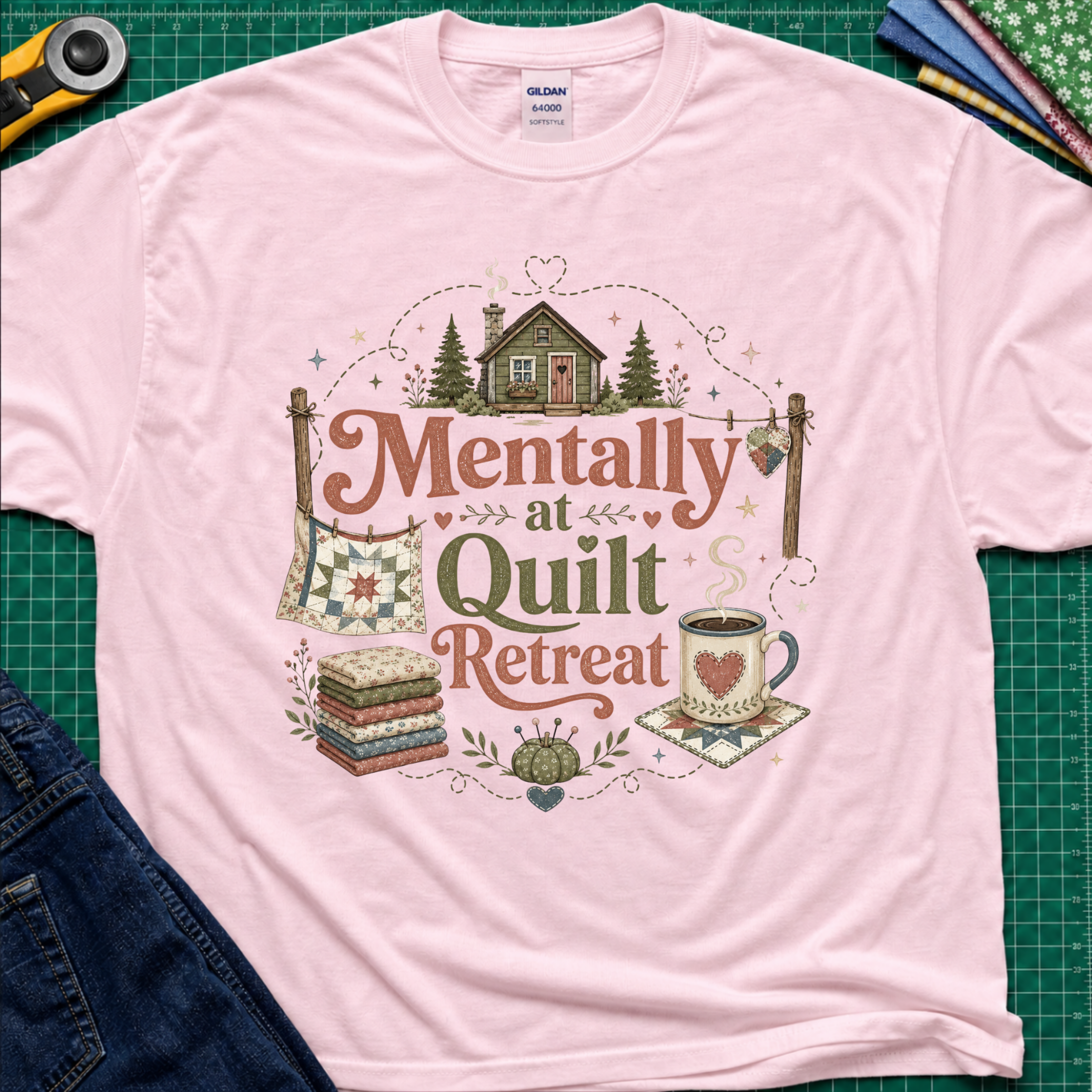 A light pink Gildan t-shirt with a rustic  style graphic reading 'Mentally at Quilt Retreat' surrounded by quilts, a cozy cabin, a coffee cup, and a stack of quilts. The shirt is styled flat on a green cutting mat with a rotary cutter, blue jeans and fabric stack.