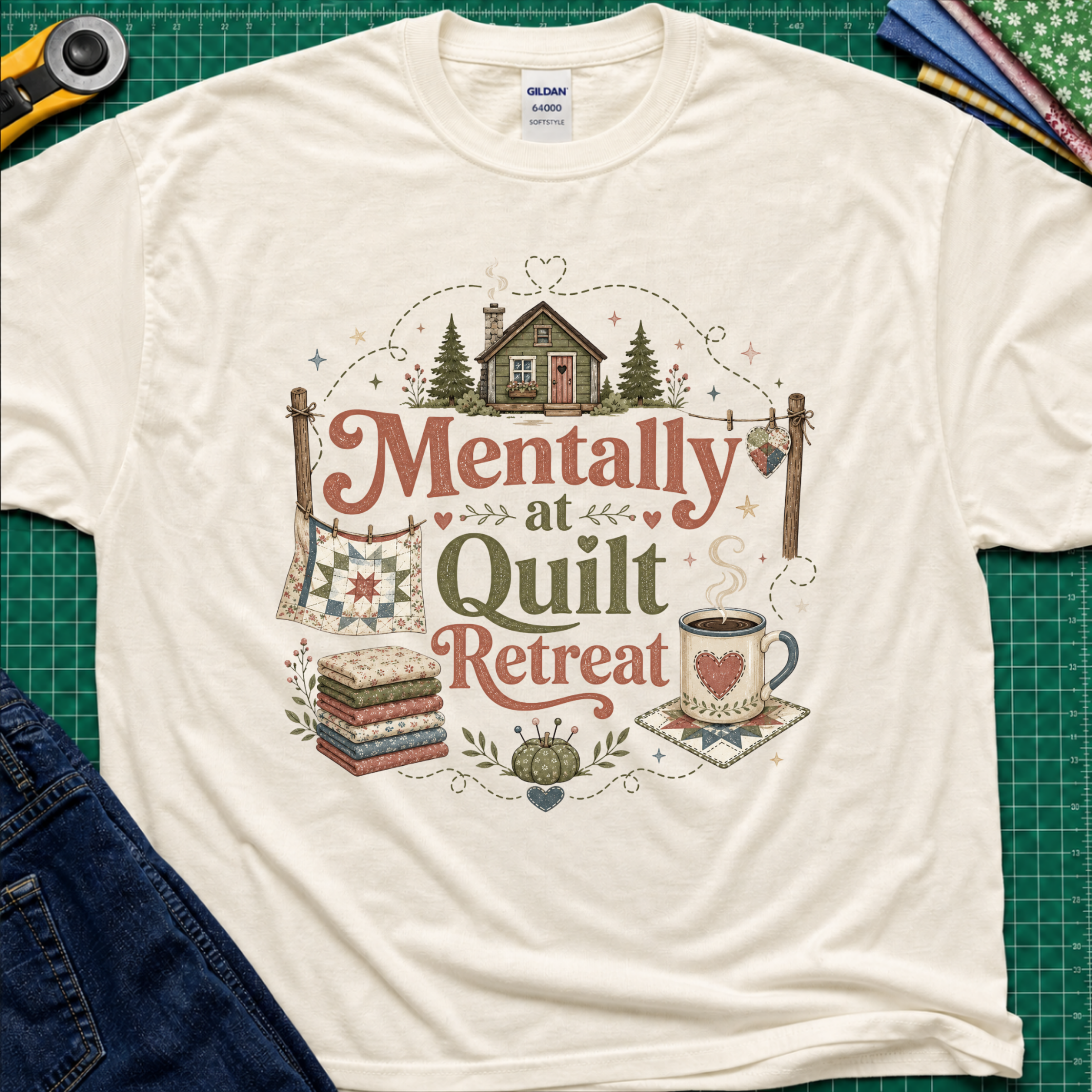 A light cream Gildan t-shirt with a rustic  style graphic reading 'Mentally at Quilt Retreat' surrounded by quilts, a cozy cabin, a coffee cup, and a stack of quilts. The shirt is styled flat on a green cutting mat with a rotary cutter, blue jeans and fabric stack.