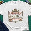 A white Gildan t-shirt with a rustic  style graphic reading 'Mentally at Quilt Retreat' surrounded by quilts, a cozy cabin, a coffee cup, and a stack of quilts. The shirt is styled flat on a green cutting mat with a rotary cutter, blue jeans and fabric stack.
