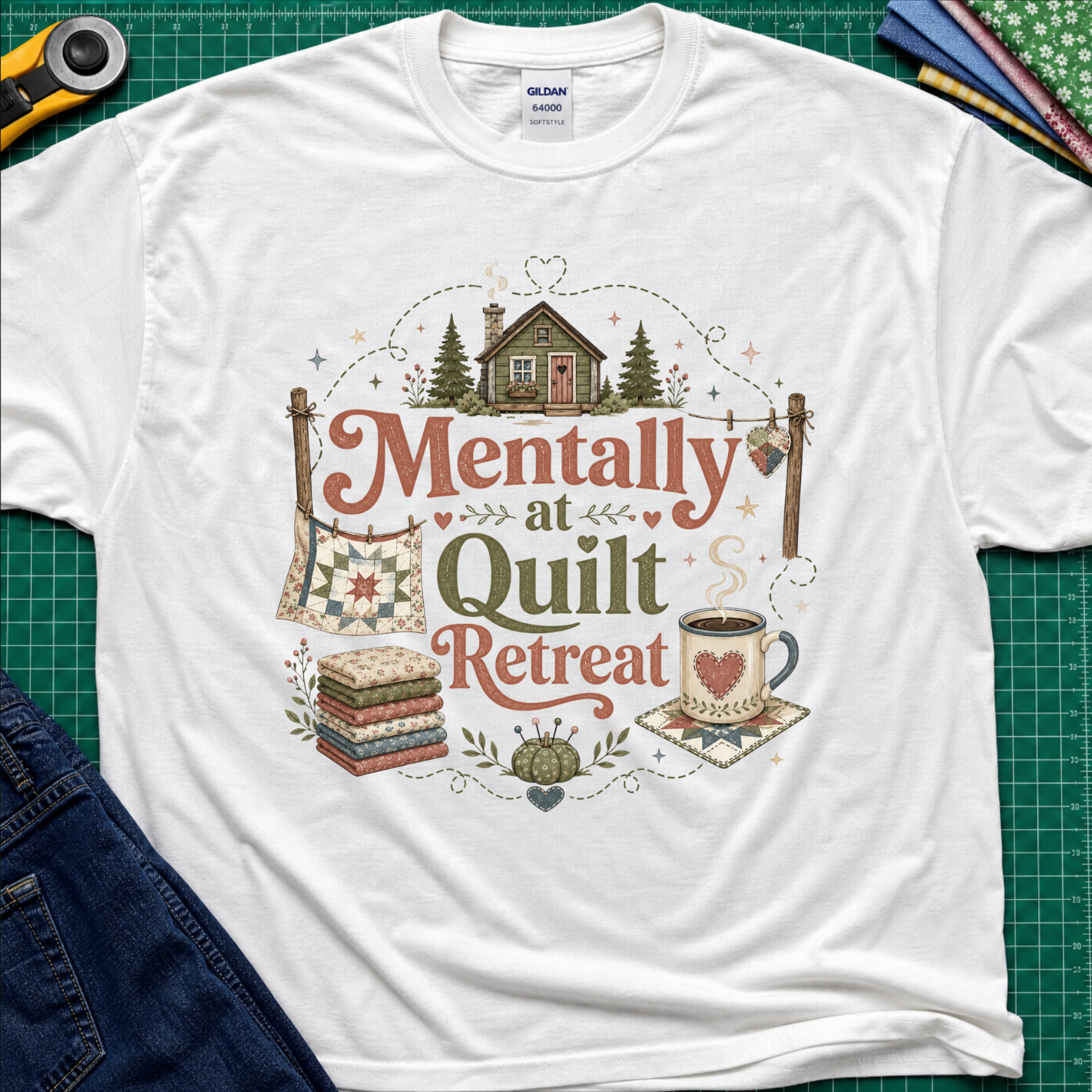 A white Gildan t-shirt with a rustic  style graphic reading 'Mentally at Quilt Retreat' surrounded by quilts, a cozy cabin, a coffee cup, and a stack of quilts. The shirt is styled flat on a green cutting mat with a rotary cutter, blue jeans and fabric stack.