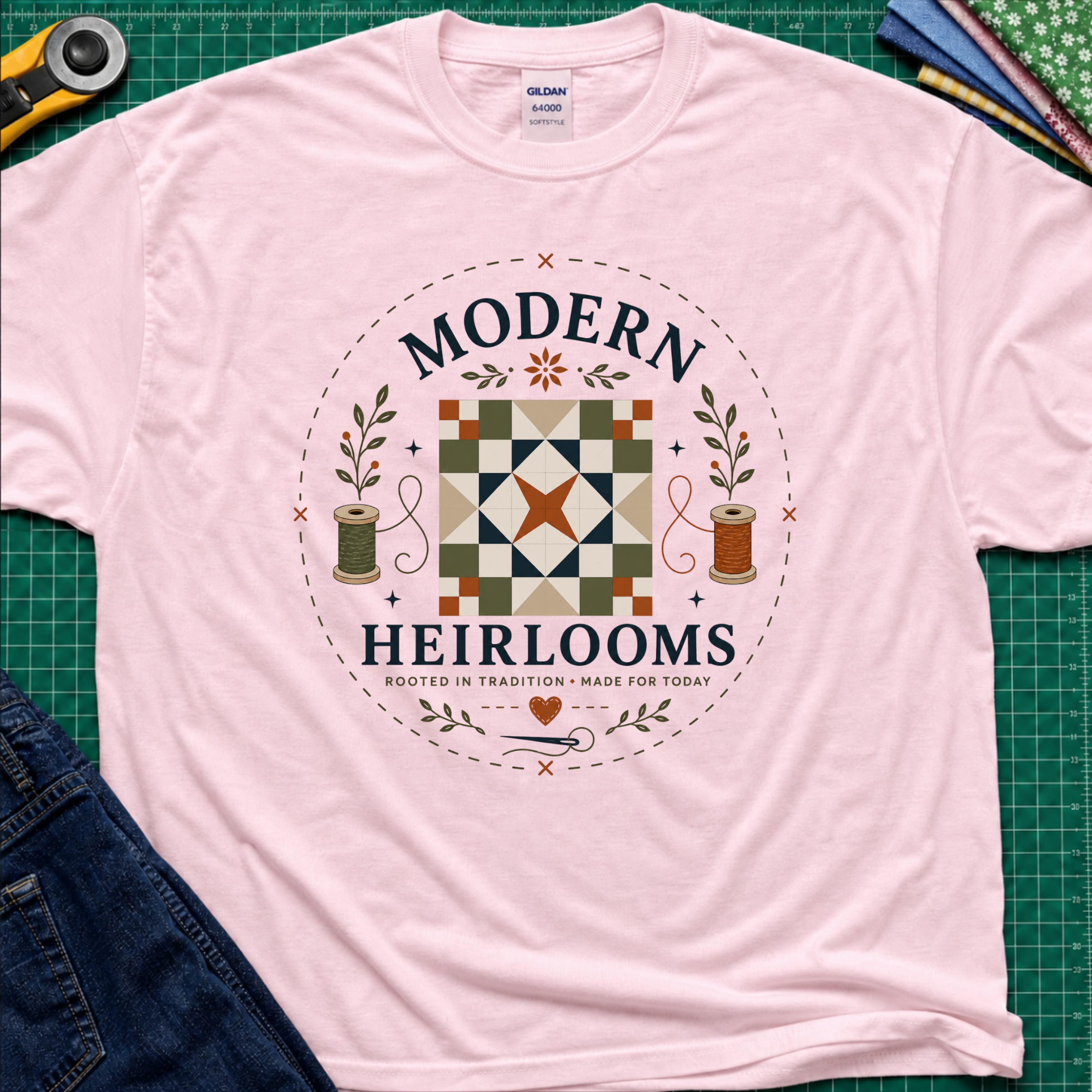 A light pink Gildan t-shirt with a circular 'Modern Heirlooms' quilt graphic around a quilt block, thread spools, needle and small leaves. Styled flat on a green cutting mat with sewing props.