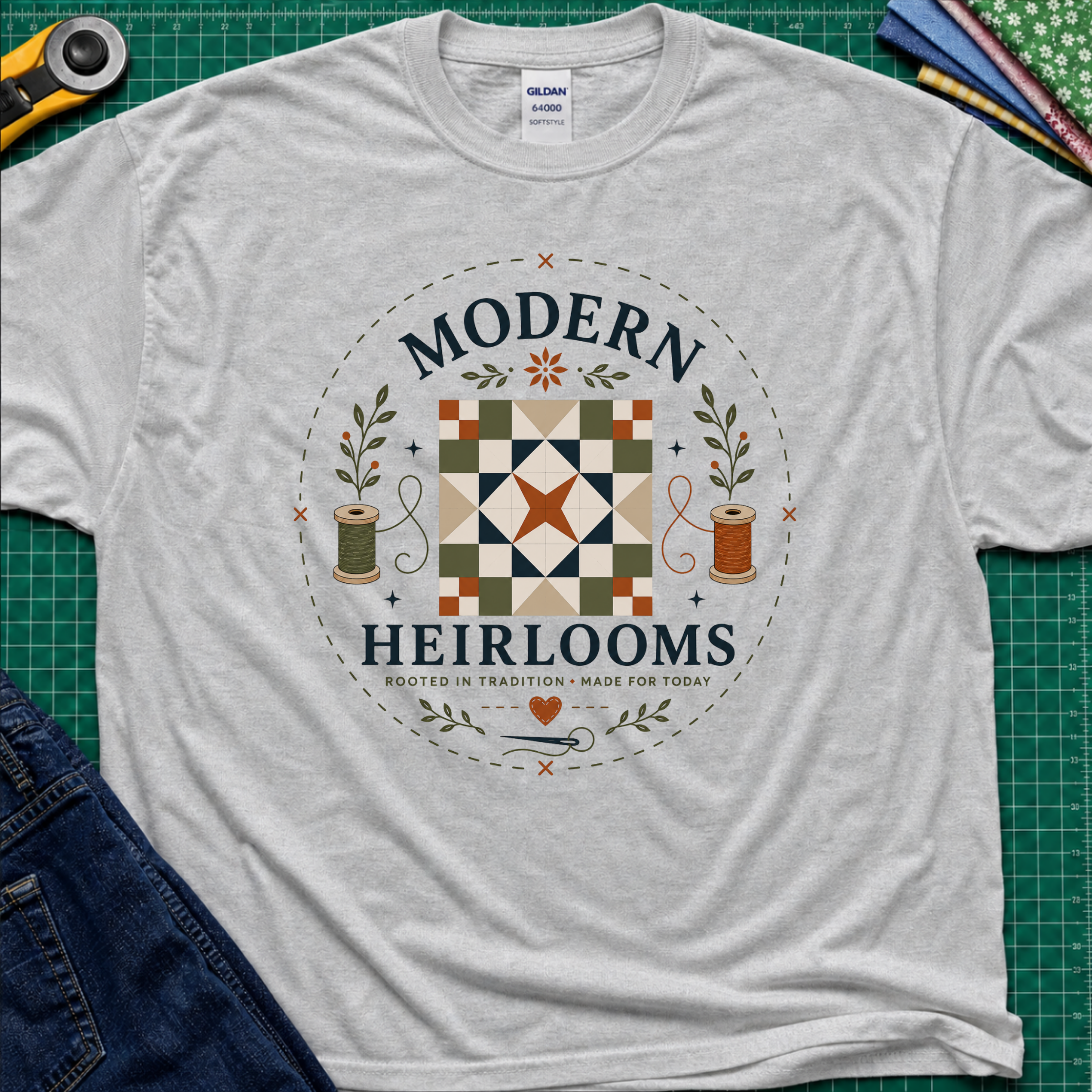 A light sports grey Gildan t-shirt with a circular 'Modern Heirlooms' quilt graphic around a quilt block, thread spools, needle and small leaves. Styled flat on a green cutting mat with sewing props.