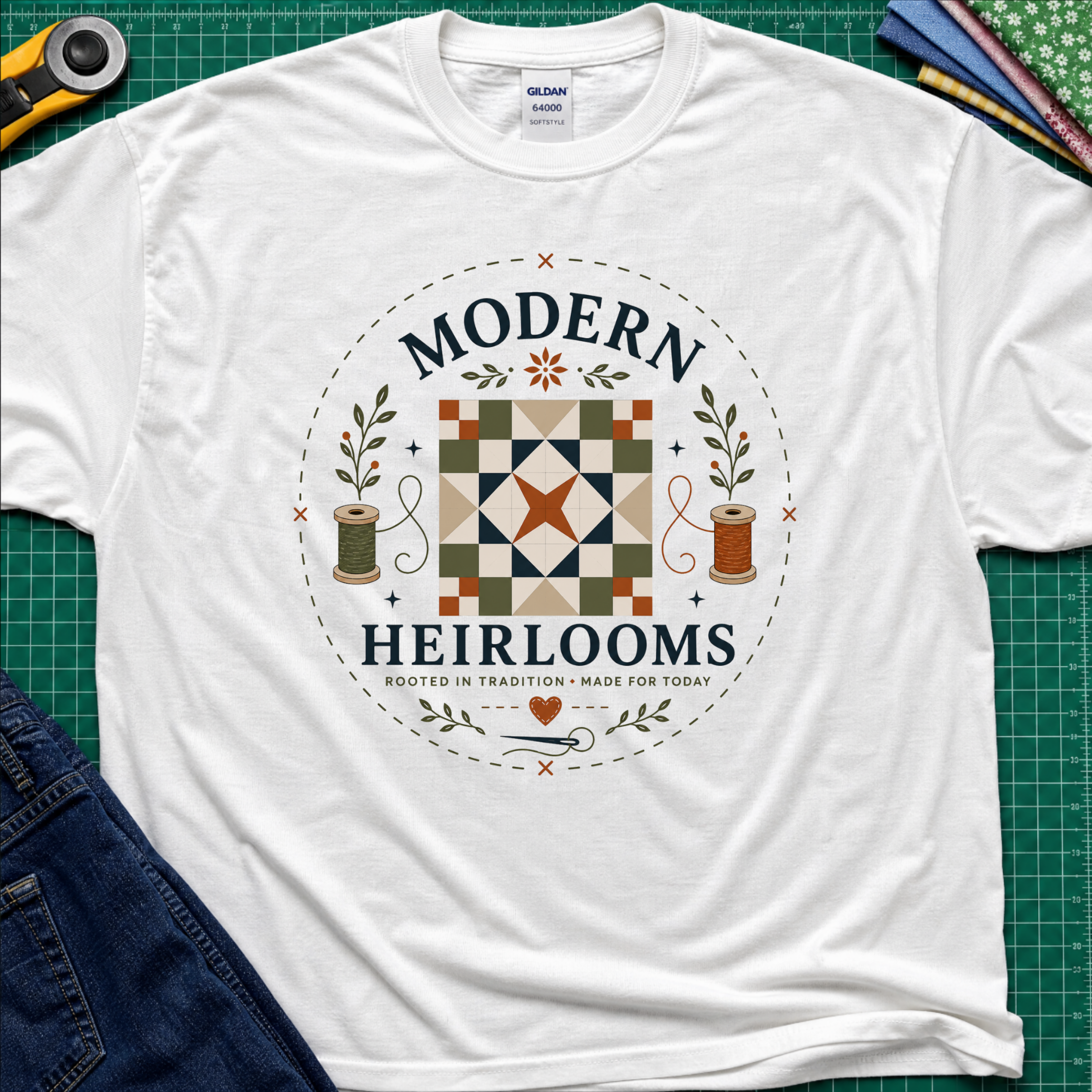 A white Gildan t-shirt with a circular 'Modern Heirlooms' quilt graphic around a quilt block, thread spools, needle and small leaves. Styled flat on a green cutting mat with sewing props.
