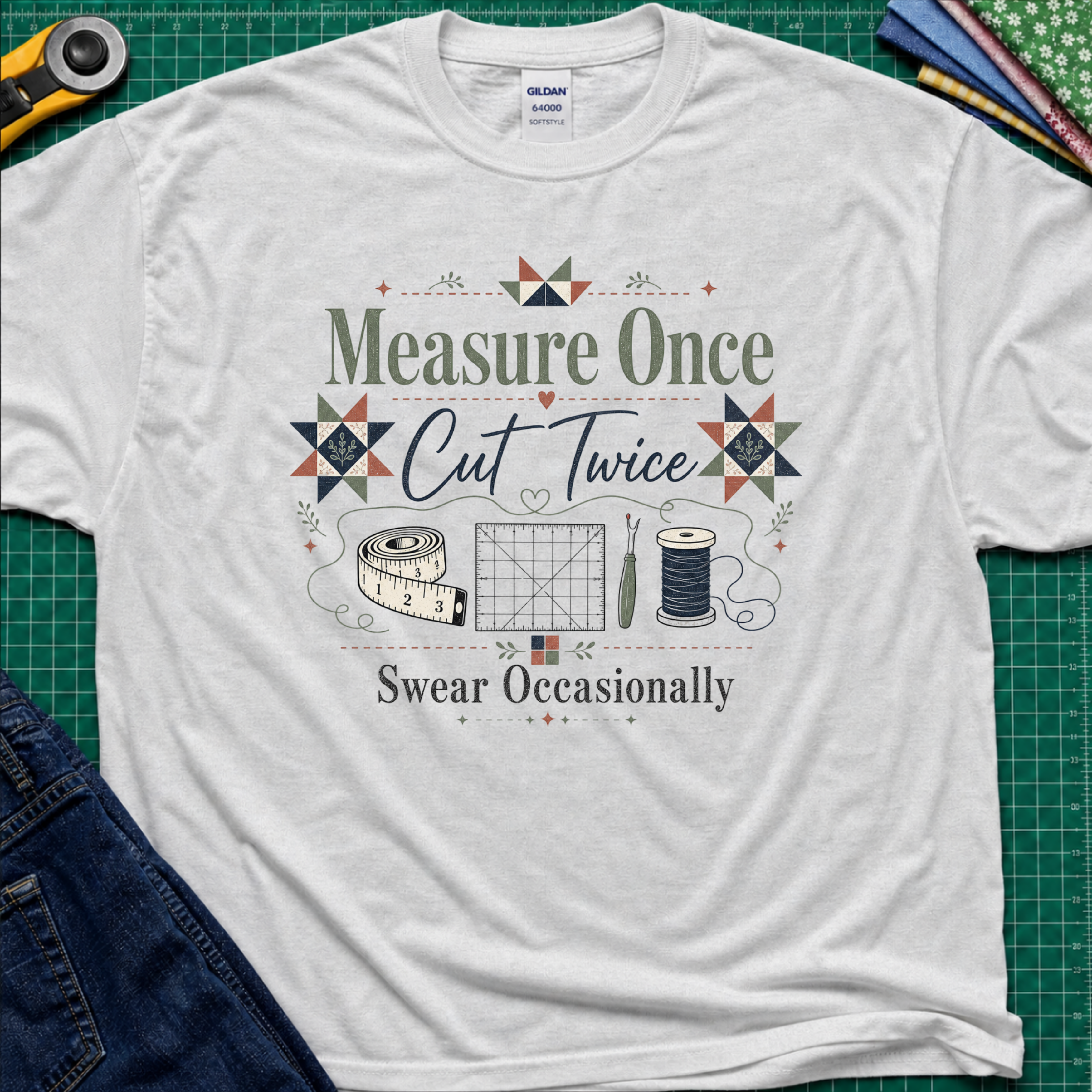 A light grey Gildan t-shirt with a vintage-style quilting graphic that reads 'Measure Once, Cut Twice, Swear Occasionally' surrounded by sewing tools and geometric patterns, laid flat on a green cutting mat with sewing props.