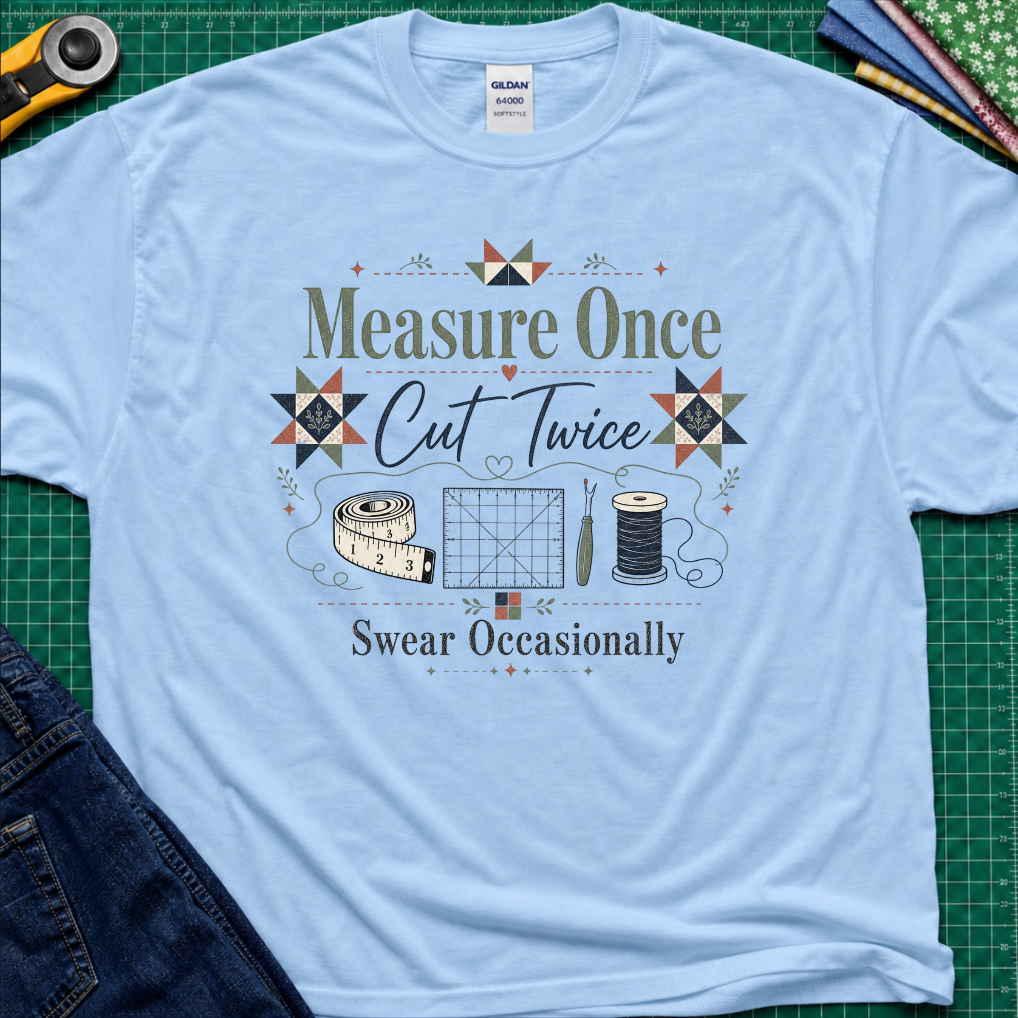 A light blue Gildan t-shirt with a vintage-style quilting graphic that reads 'Measure Once, Cut Twice, Swear Occasionally' surrounded by sewing tools and geometric patterns, laid flat on a green cutting mat with sewing props.