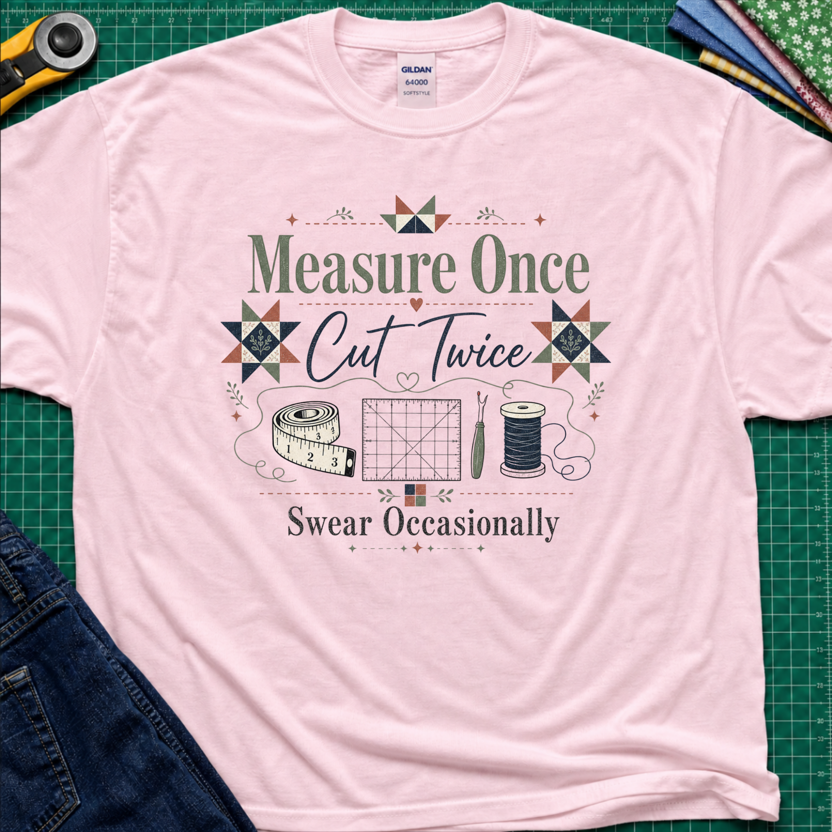 A light pink Gildan t-shirt with a vintage-style quilting graphic that reads 'Measure Once, Cut Twice, Swear Occasionally' surrounded by sewing tools and geometric patterns, laid flat on a green cutting mat with sewing props.