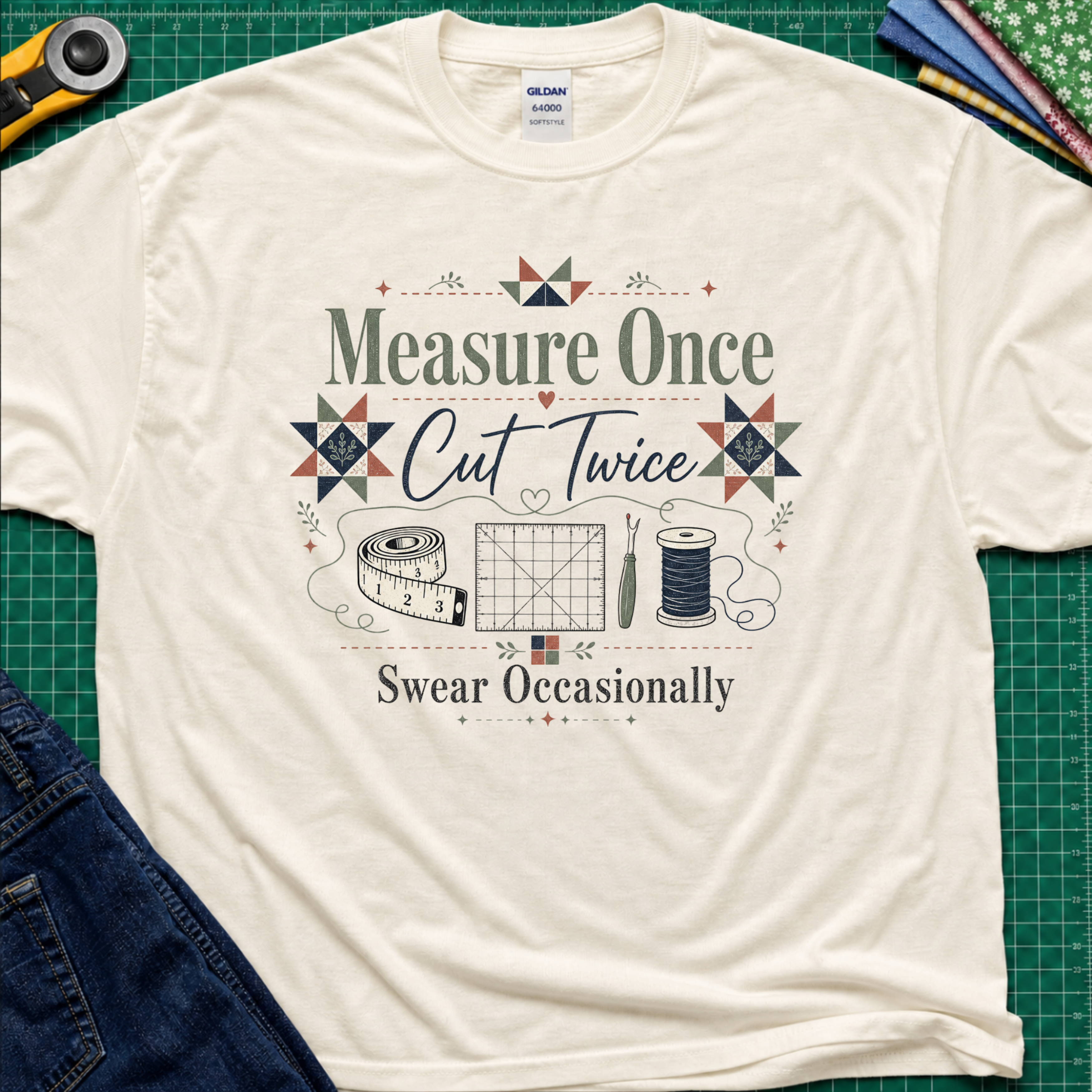 A light cream Gildan t-shirt with a vintage-style quilting graphic that reads 'Measure Once, Cut Twice, Swear Occasionally' surrounded by sewing tools and geometric patterns, laid flat on a green cutting mat with sewing props.