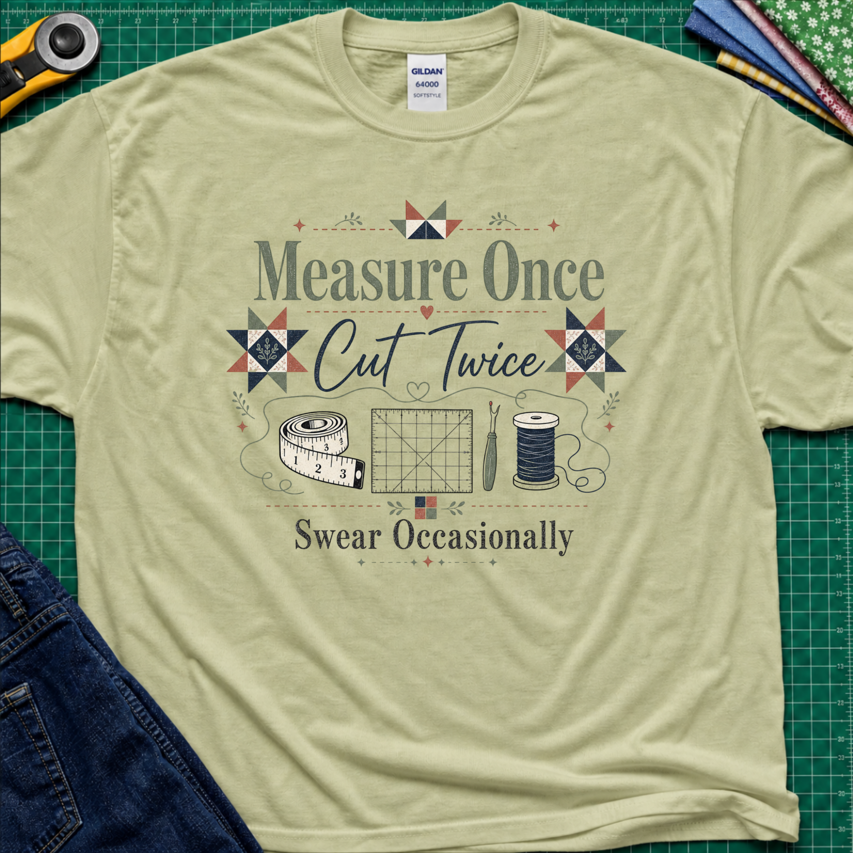 A light pistachio green Gildan t-shirt with a vintage-style quilting graphic that reads 'Measure Once, Cut Twice, Swear Occasionally' surrounded by sewing tools and geometric patterns, laid flat on a green cutting mat with sewing props.