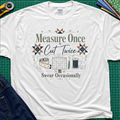 A white Gildan t-shirt with a vintage-style quilting graphic that reads 'Measure Once, Cut Twice, Swear Occasionally' surrounded by sewing tools and geometric patterns, laid flat on a green cutting mat with sewing props.