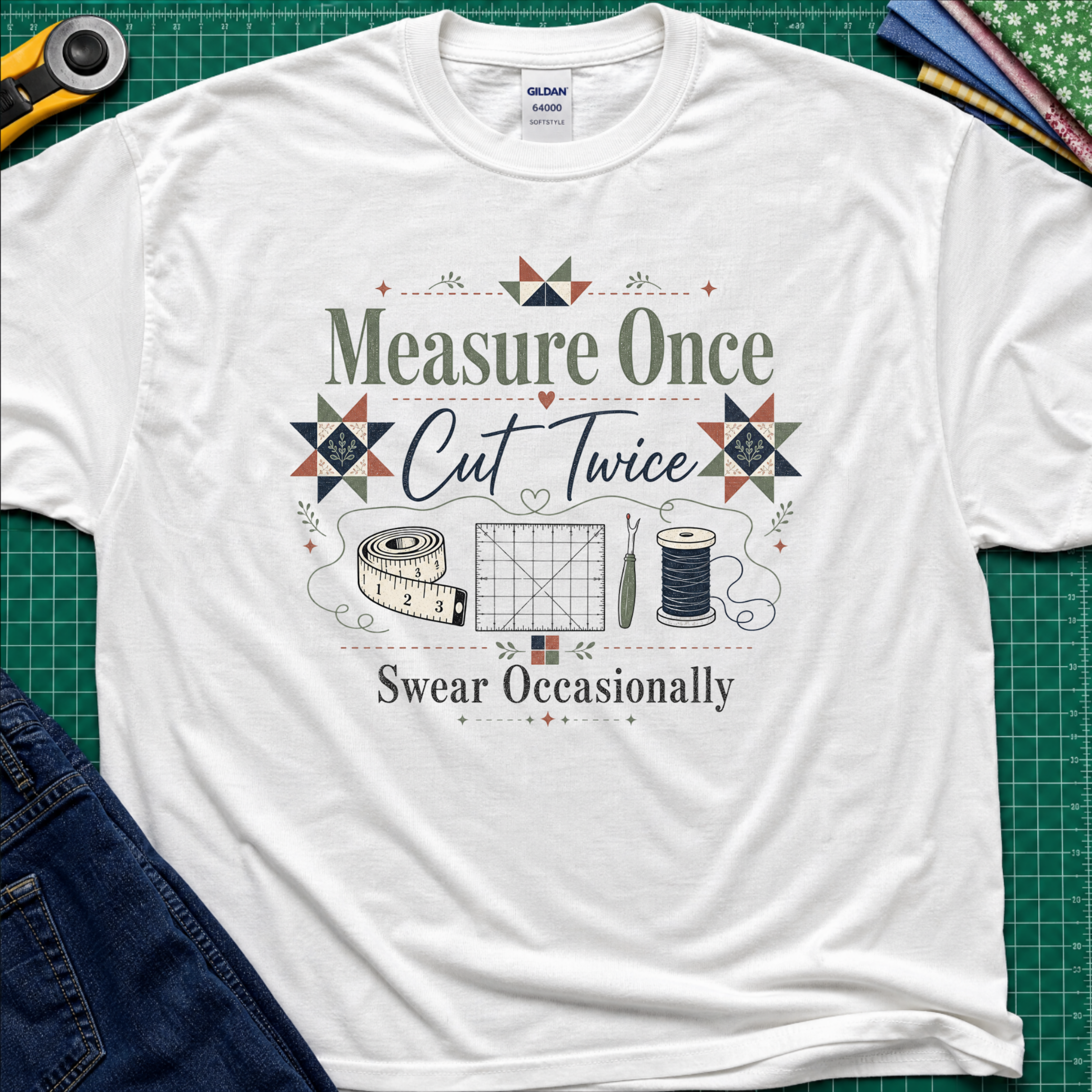 A white Gildan t-shirt with a vintage-style quilting graphic that reads 'Measure Once, Cut Twice, Swear Occasionally' surrounded by sewing tools and geometric patterns, laid flat on a green cutting mat with sewing props.
