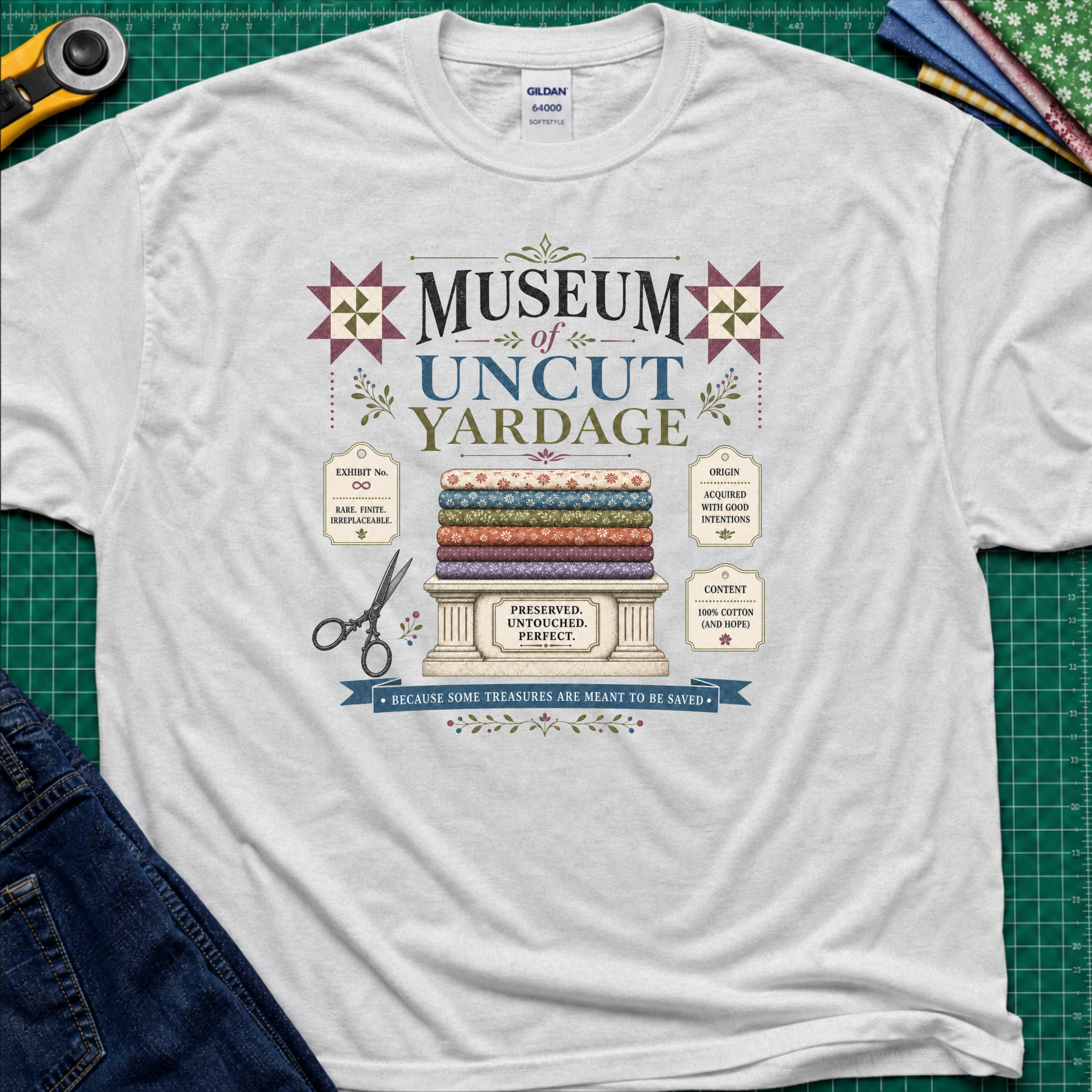 A light grey Gildan t-shirt with a vintage-style graphic reading 'Museum of Uncut Yardage' and showing stacked fabric bolts, scissors, and fabric labels. The shirt lays flat on a green cutting mat with rotary cutters, fat quarters and blue jeans partially visible in the corners.