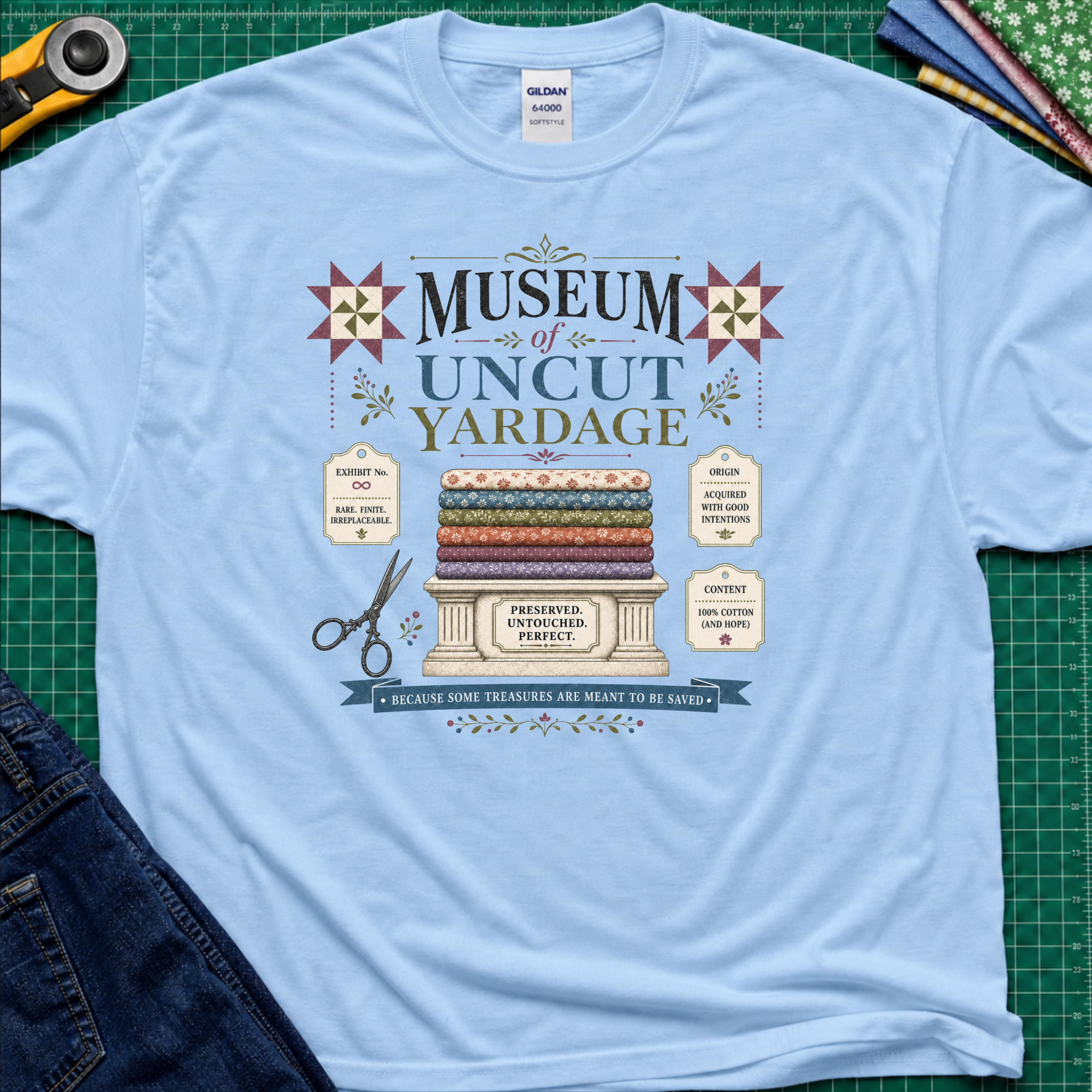 A light blue Gildan t-shirt with a vintage-style graphic reading 'Museum of Uncut Yardage' and showing stacked fabric bolts, scissors, and fabric labels. The shirt lays flat on a green cutting mat with rotary cutters, fat quarters and blue jeans partially visible in the corners.