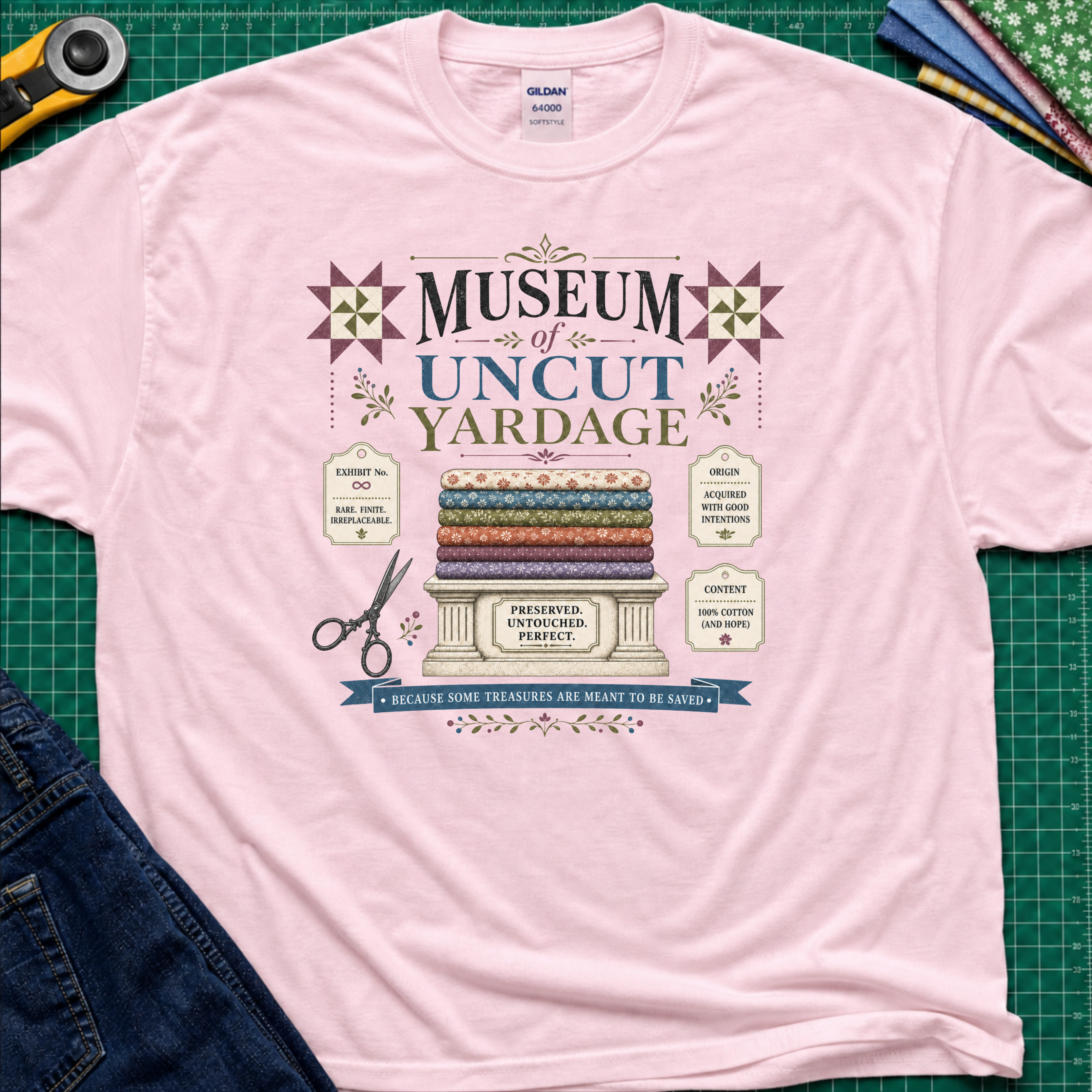 A light pink Gildan t-shirt with a vintage-style graphic reading 'Museum of Uncut Yardage' and showing stacked fabric bolts, scissors, and fabric labels. The shirt lays flat on a green cutting mat with rotary cutters, fat quarters and blue jeans partially visible in the corners.