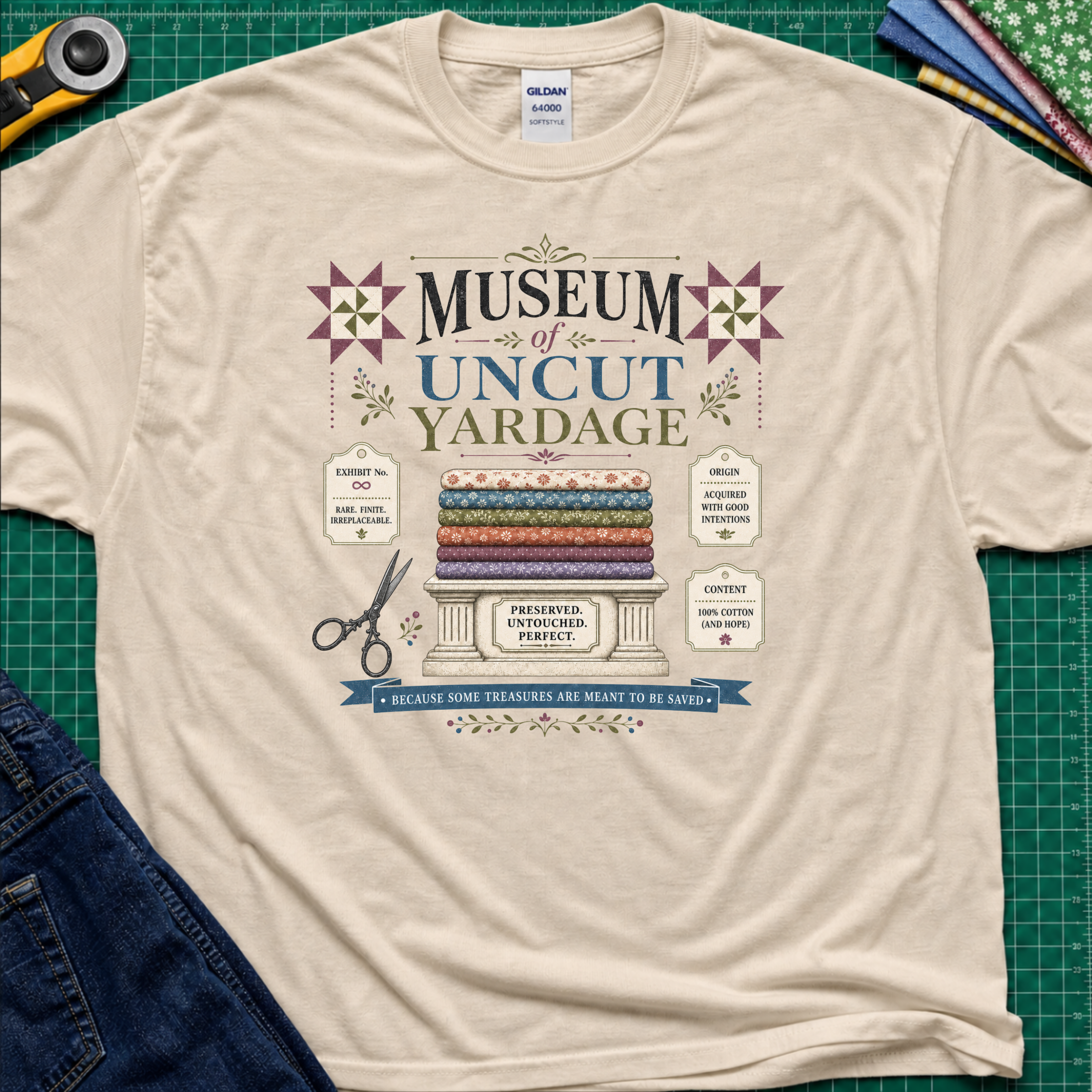 A dark cream Gildan t-shirt with a vintage-style graphic reading 'Museum of Uncut Yardage' and showing stacked fabric bolts, scissors, and fabric labels. The shirt lays flat on a green cutting mat with rotary cutters, fat quarters and blue jeans partially visible in the corners.