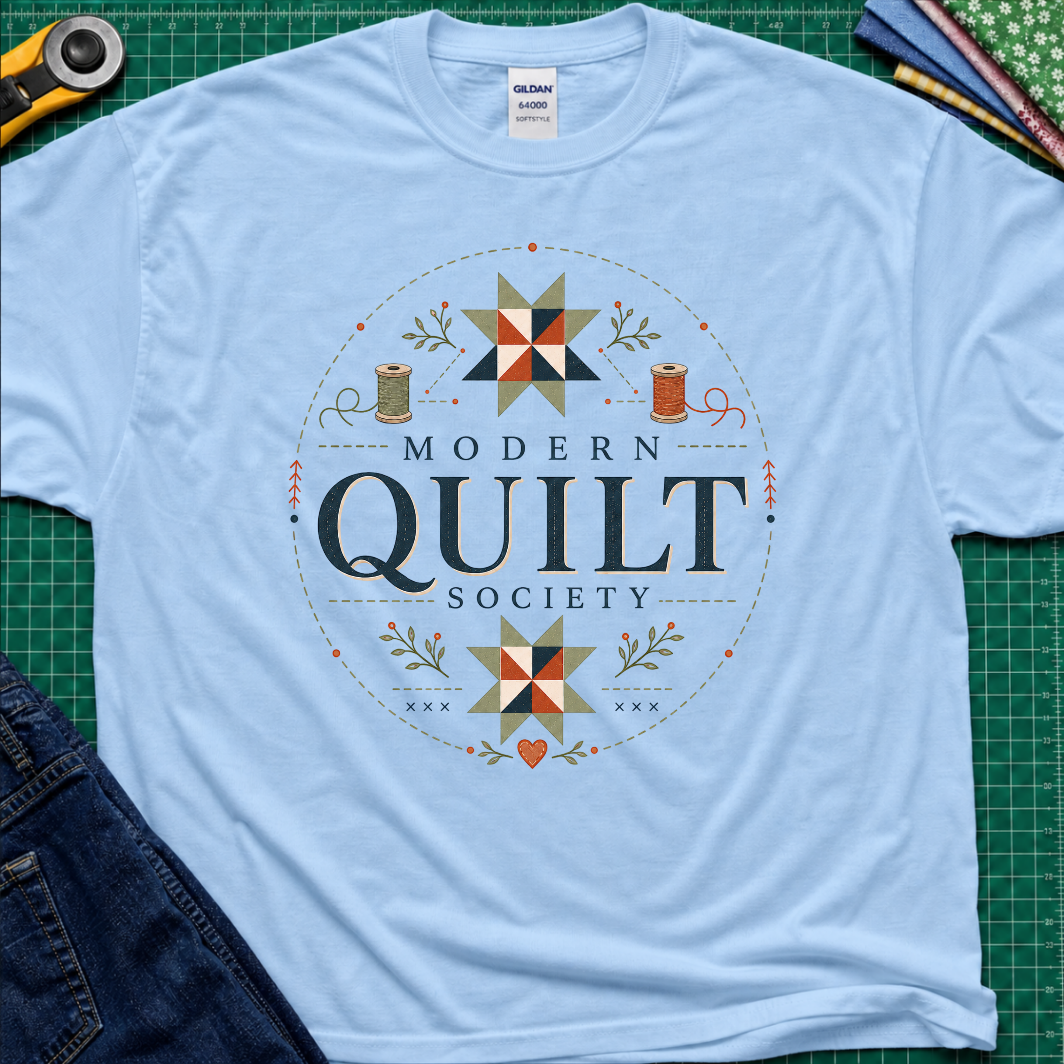 A light blue Gildan t-shirt with a 'Modern Quilt Society' graphic printed on the front, with quilt blocks, thread spools and small leaves. Styled in a flat-lay on a green cutting mat with sewing tools and fabric.