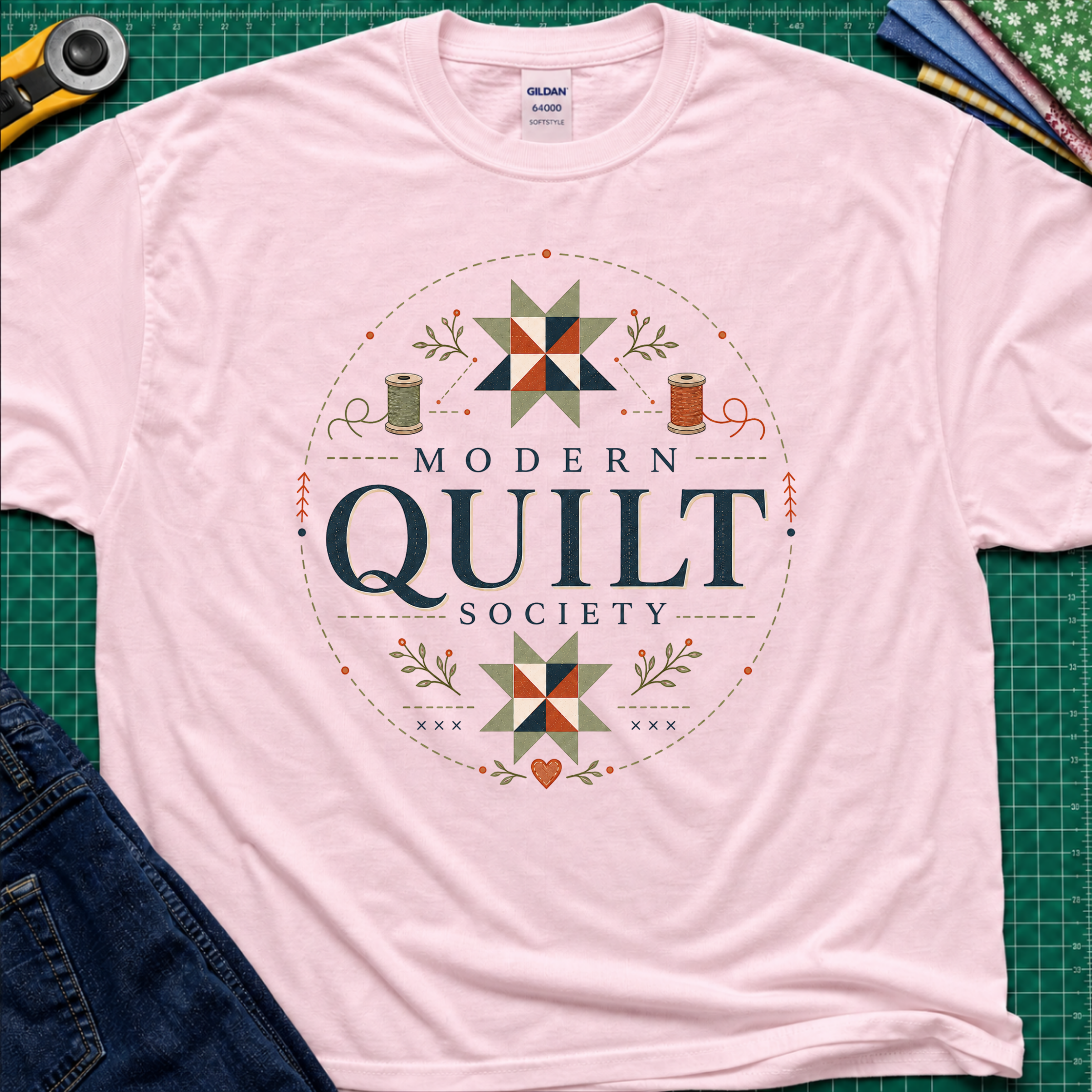 A light pink Gildan t-shirt with a 'Modern Quilt Society' graphic printed on the front, with quilt blocks, thread spools and small leaves. Styled in a flat-lay on a green cutting mat with sewing tools and fabric.