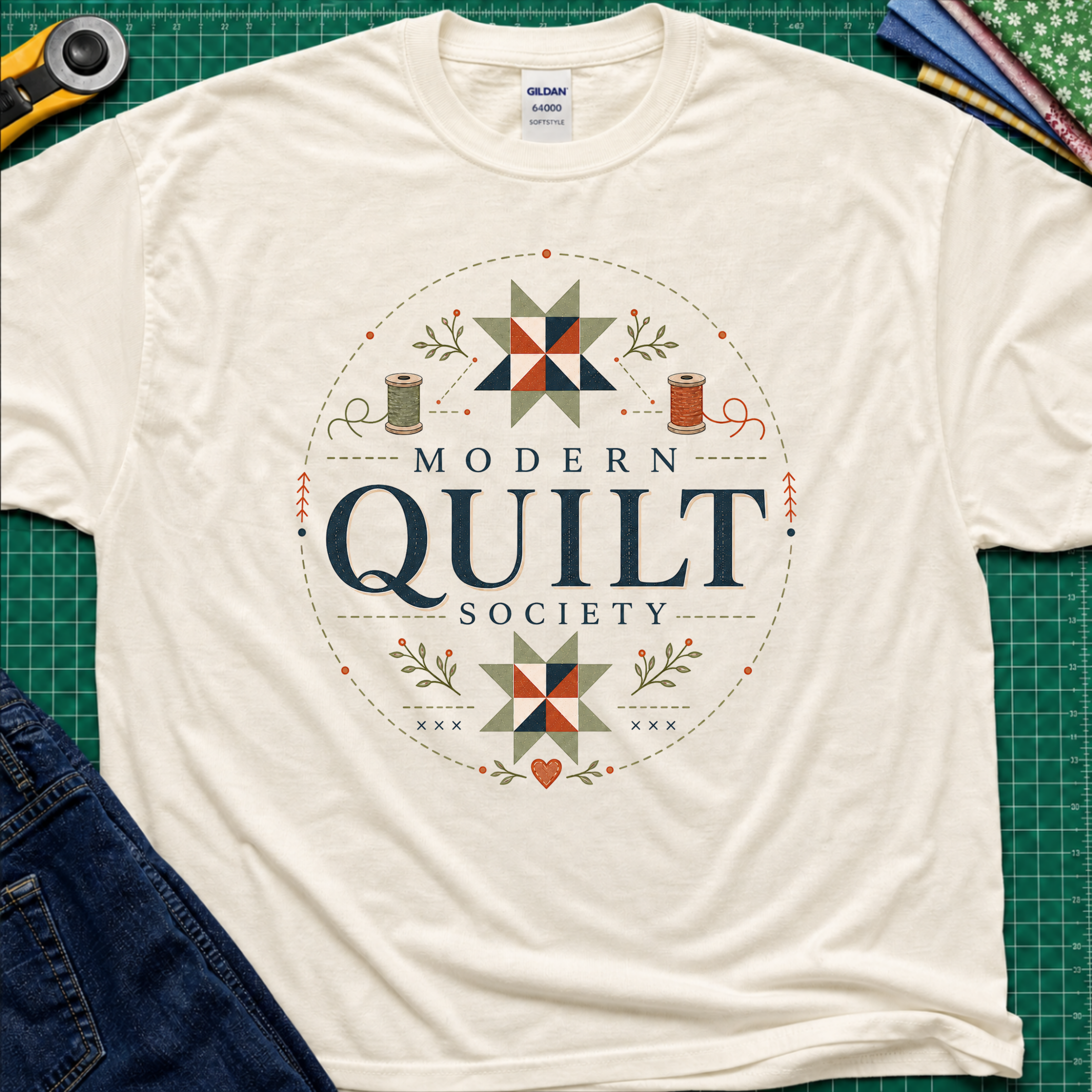 A light cream Gildan t-shirt with a 'Modern Quilt Society' graphic printed on the front, with quilt blocks, thread spools and small leaves. Styled in a flat-lay on a green cutting mat with sewing tools and fabric.
