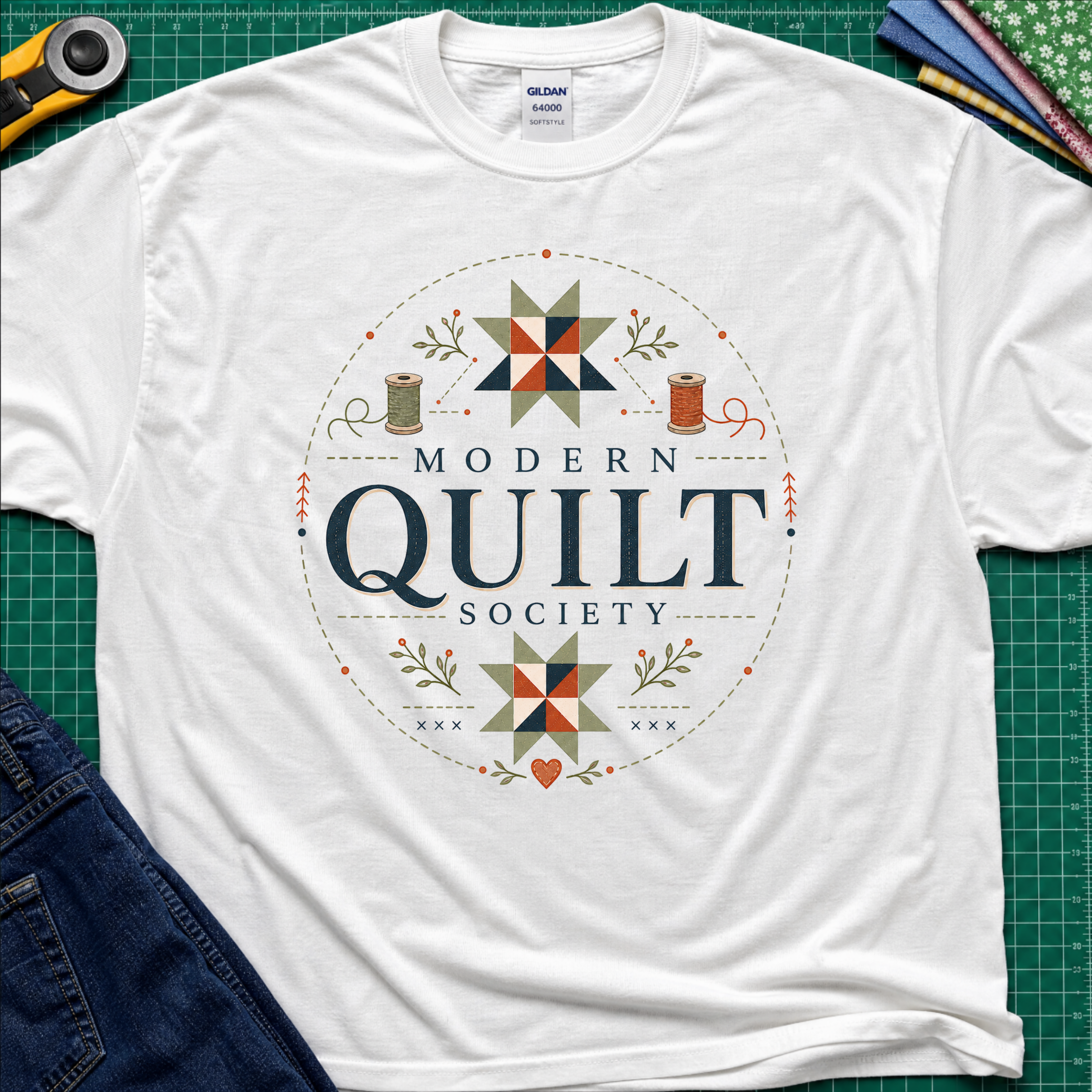 A white Gildan t-shirt with a 'Modern Quilt Society' graphic printed on the front, with quilt blocks, thread spools and small leaves. Styled in a flat-lay on a green cutting mat with sewing tools and fabric.