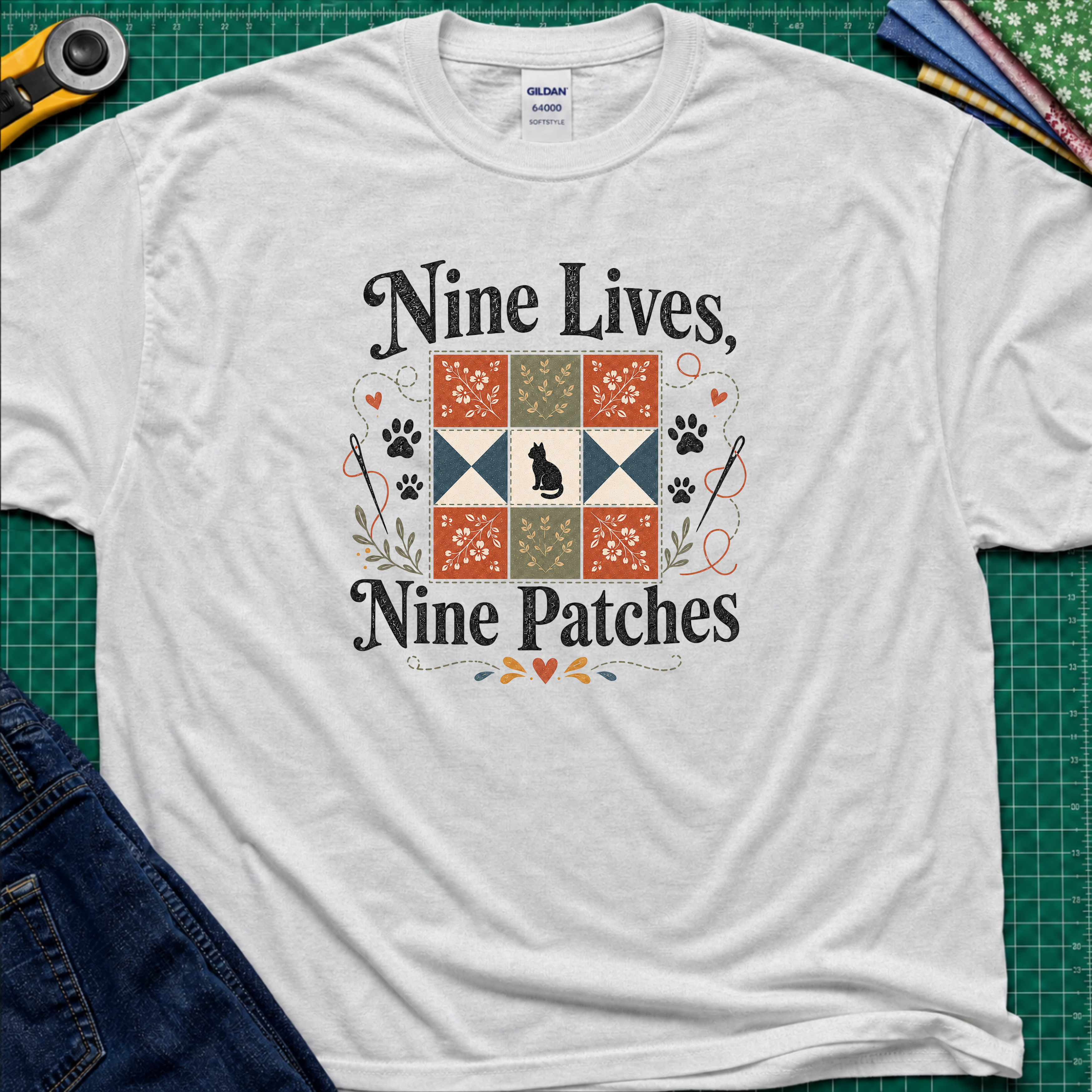 A light grey Gildan t-shirt laid flat on a green cutting mat, featuring a black applique cat centred in a nine patch block with the text 'Nine Lives, Nine Patches'. Rotary cutter, fabric stack and blue jeans surround the shirt.