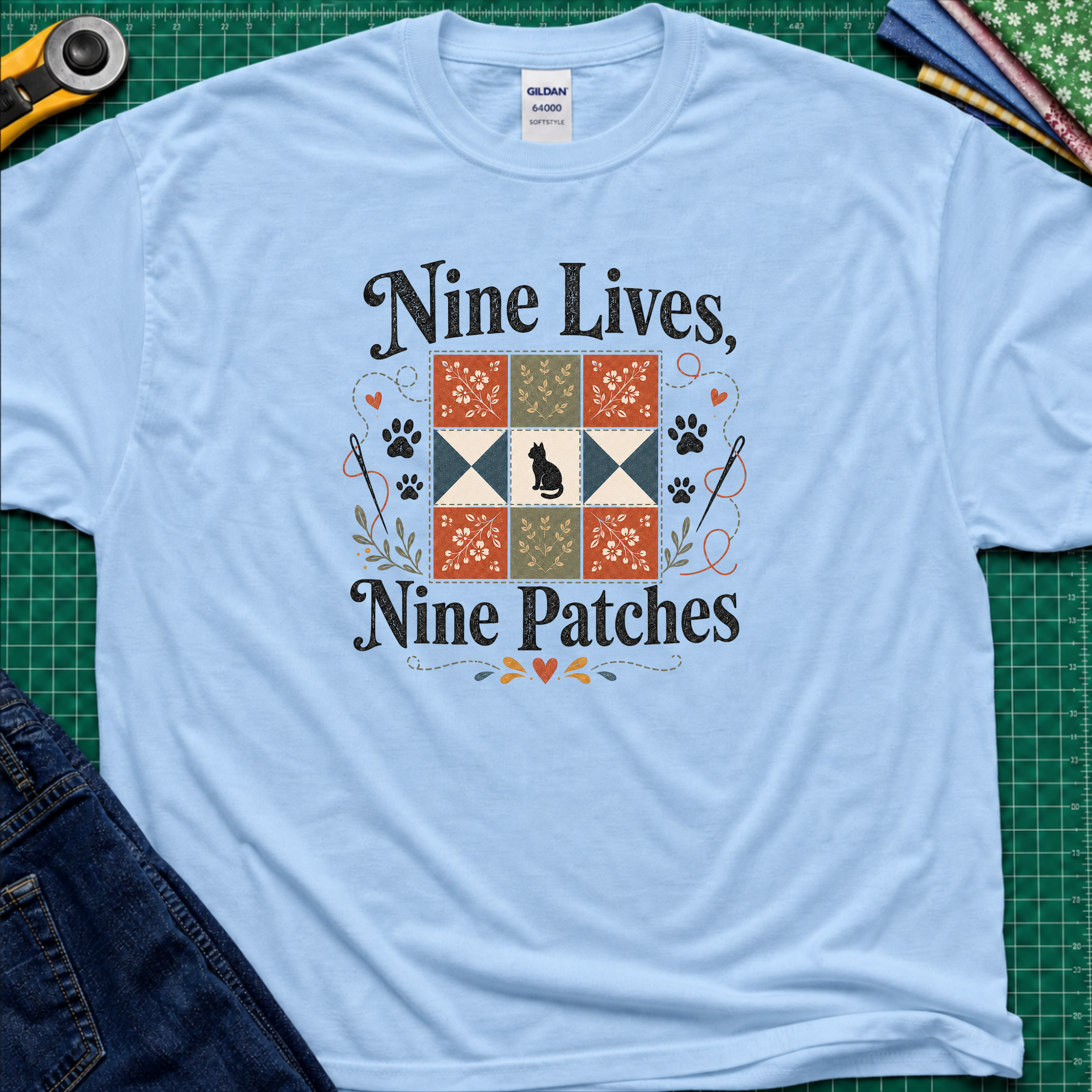 A light blue Gildan t-shirt laid flat on a green cutting mat, featuring a black applique cat centred in a nine patch block with the text 'Nine Lives, Nine Patches'. Rotary cutter, fabric stack and blue jeans surround the shirt.