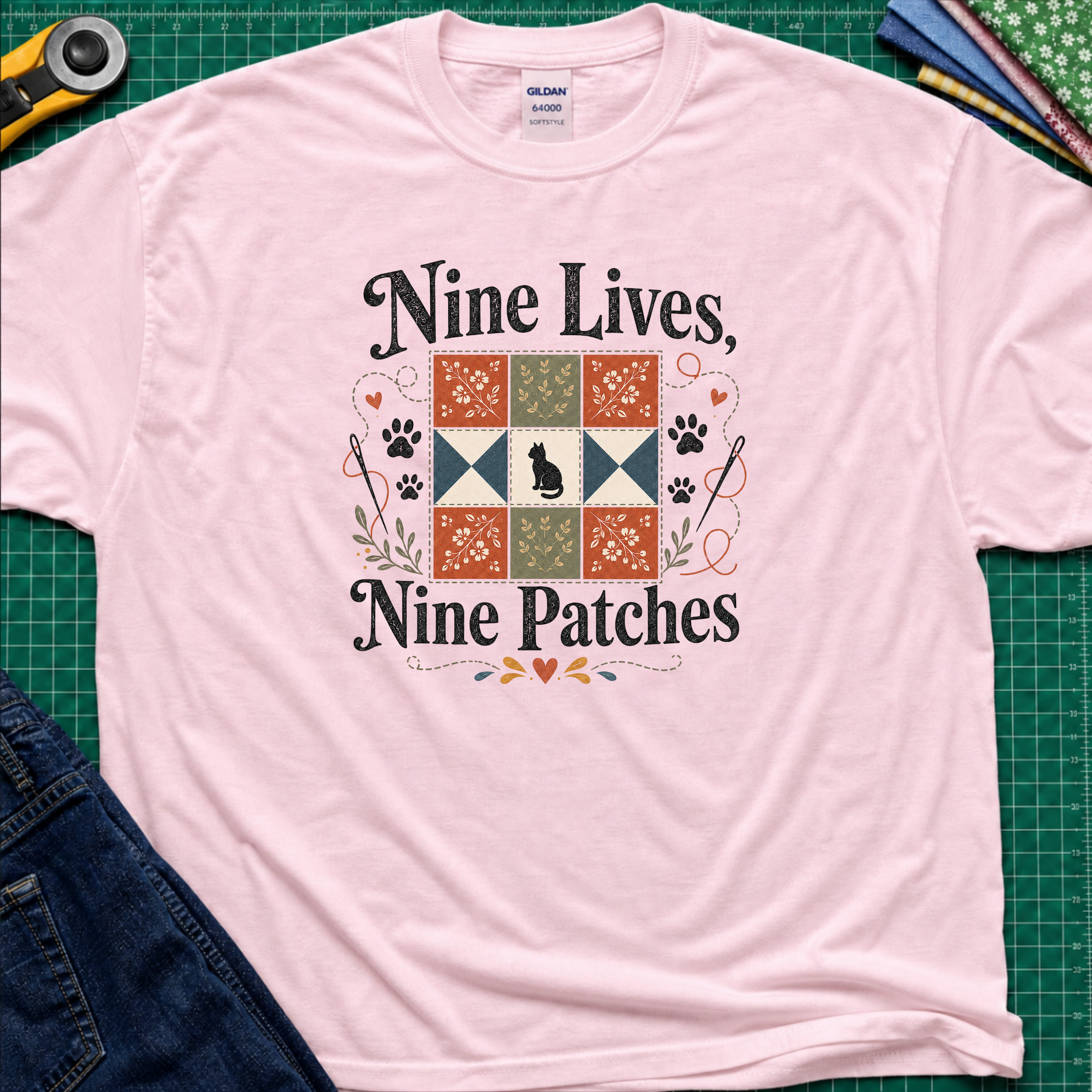 A light pink Gildan t-shirt laid flat on a green cutting mat, featuring a black applique cat centred in a nine patch block with the text 'Nine Lives, Nine Patches'. Rotary cutter, fabric stack and blue jeans surround the shirt.