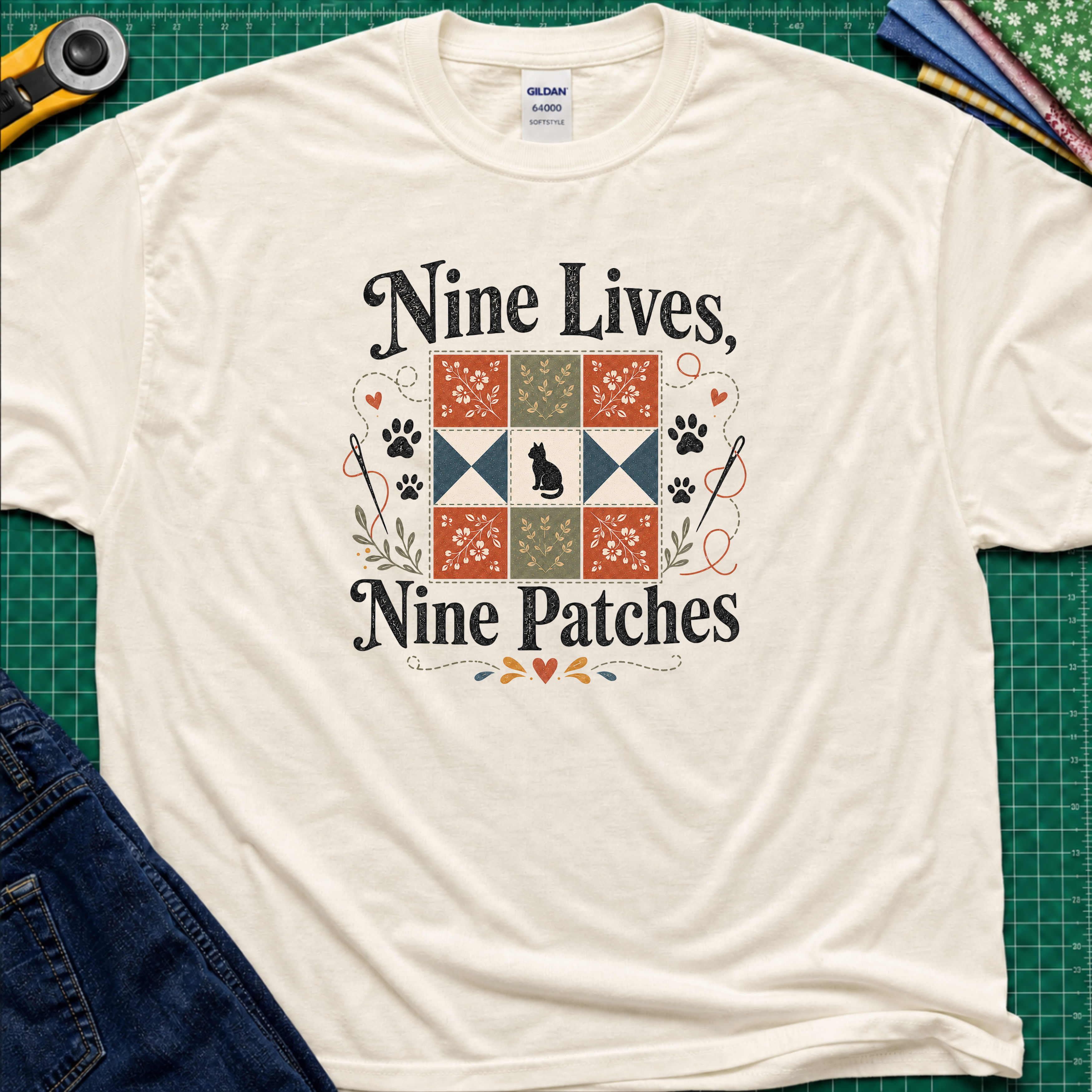 A light cream Gildan t-shirt laid flat on a green cutting mat, featuring a black applique cat centred in a nine patch block with the text 'Nine Lives, Nine Patches'. Rotary cutter, fabric stack and blue jeans surround the shirt.