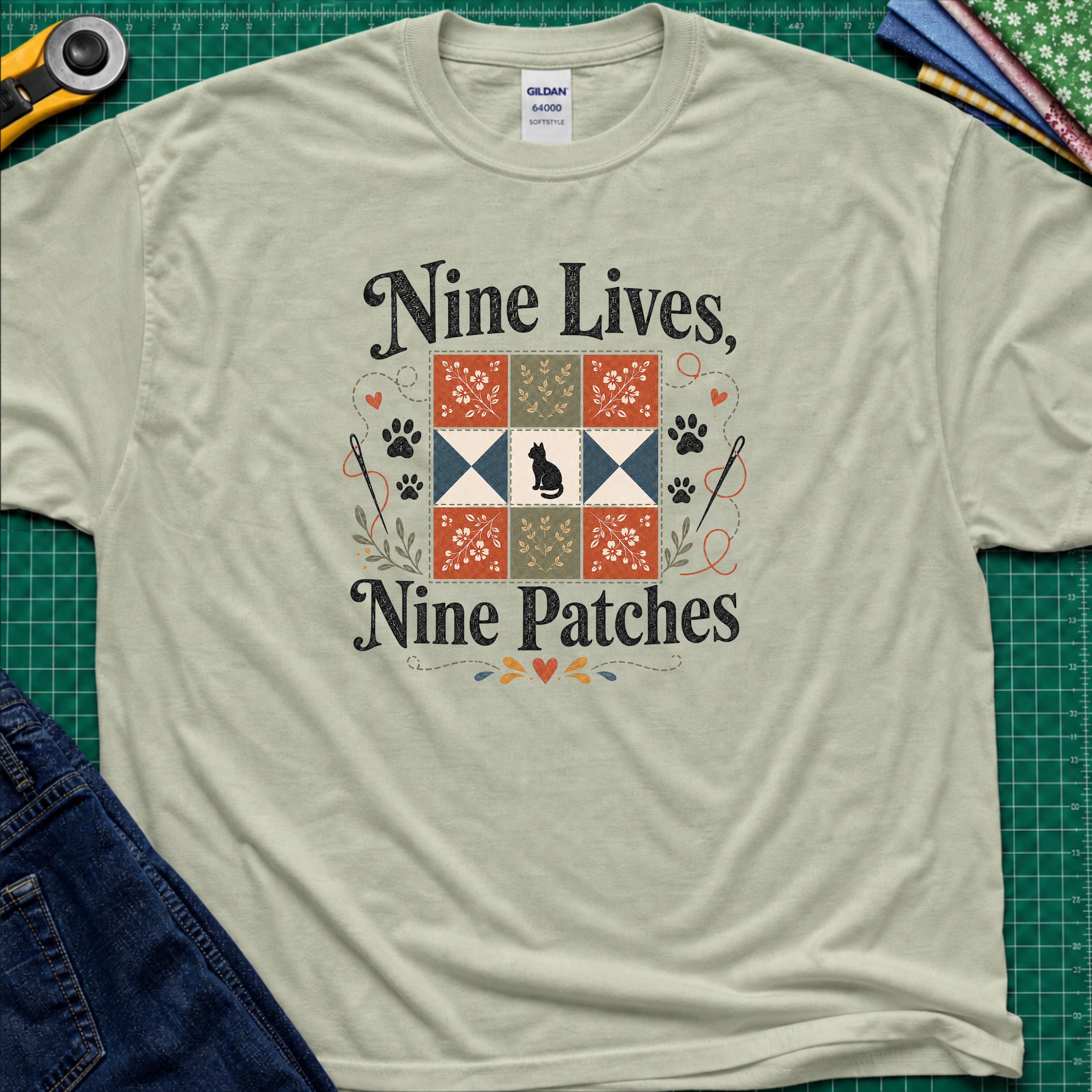 A light sage green Gildan t-shirt laid flat on a green cutting mat, featuring a black applique cat centred in a nine patch block with the text 'Nine Lives, Nine Patches'. Rotary cutter, fabric stack and blue jeans surround the shirt.