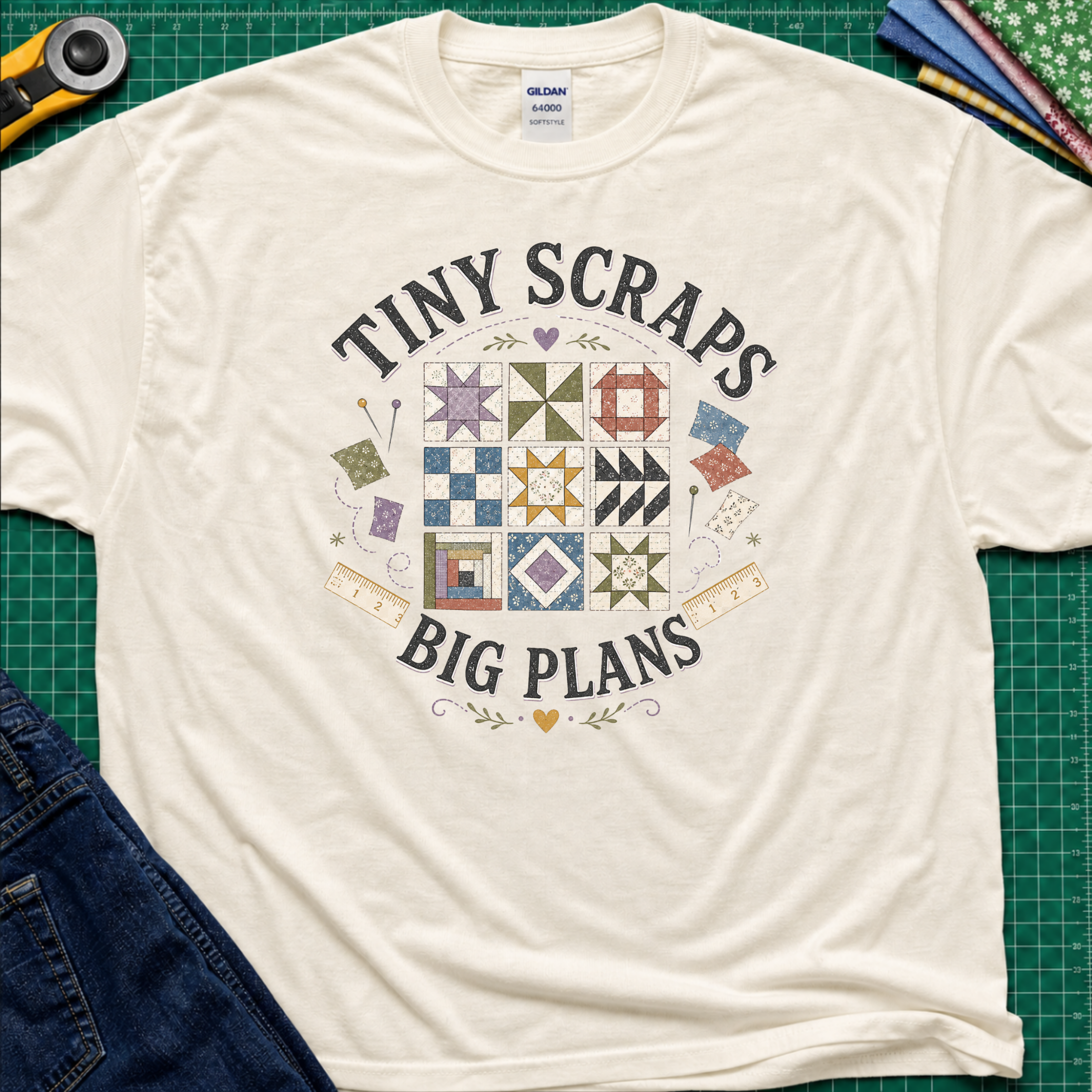 A light cream Gildan t-shirt laid flat on a green cutting mat background, featuring a quilt pattern graphic and the text 'Tiny Scraps Big Plans'. Quilting props like a rotary cutter, jeans, and fabric are partially visible around the shirt.