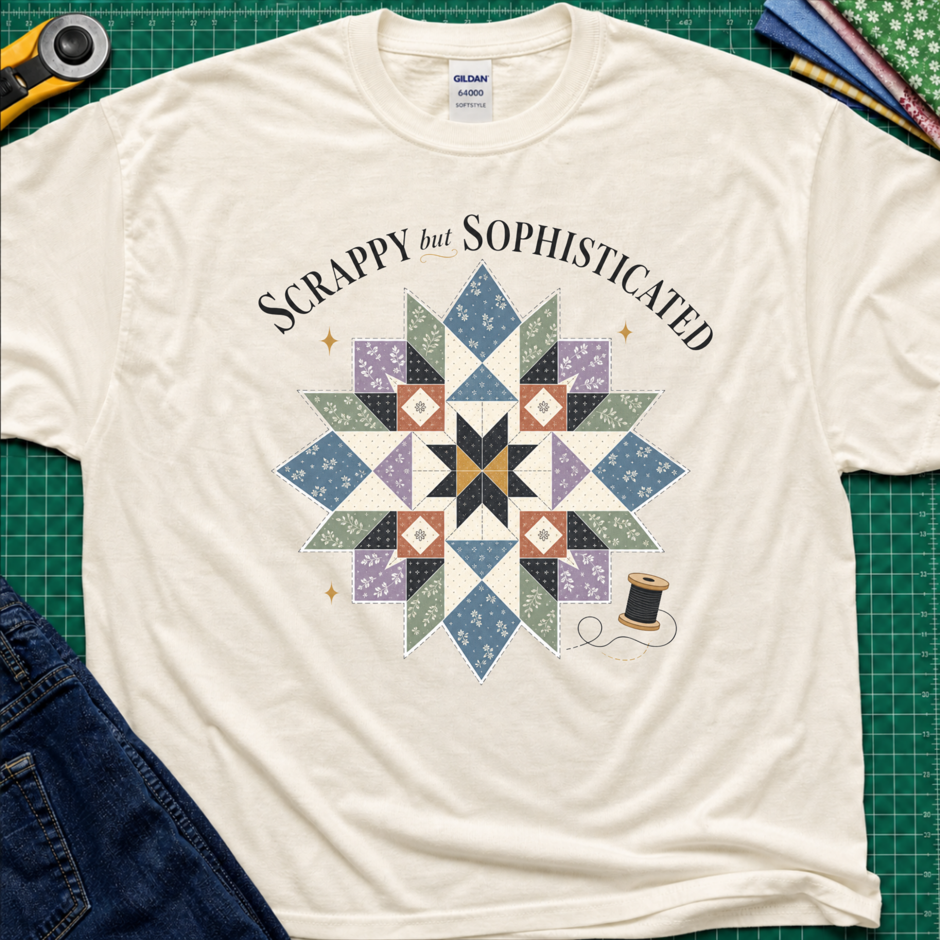 A light cream Gildan t-shirt with a colorful quilted starburst graphic and the text 'Scrappy but Sophisticated' printed on the front, styled on a green cutting mat with sewing tools and denim.