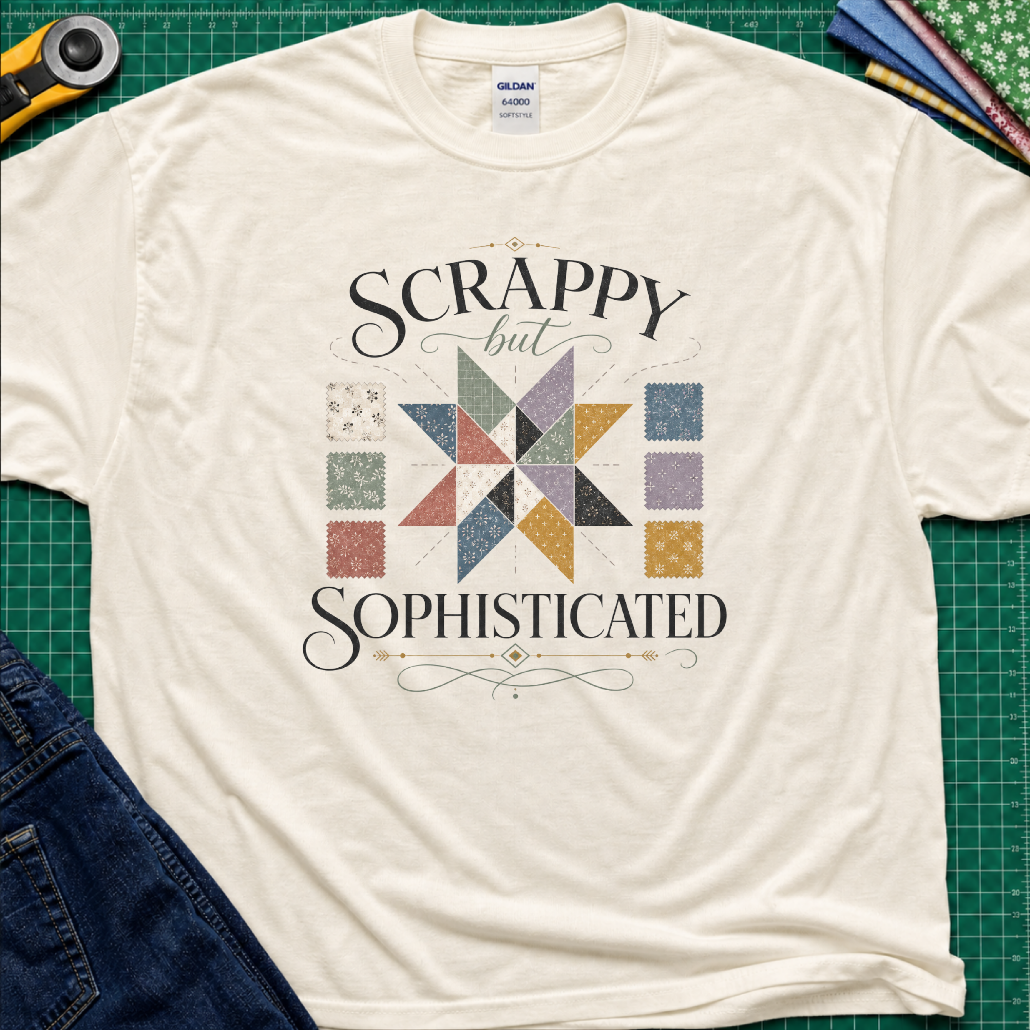 A cream Gildan t-shirt laid flat on a green cutting mat background, featuring a colorful quilted star graphic and the words 'SCRAPPY but SOPHISTICATED' in black script. A rotary cutter, fabric, and jeans are partially visible as styling props.