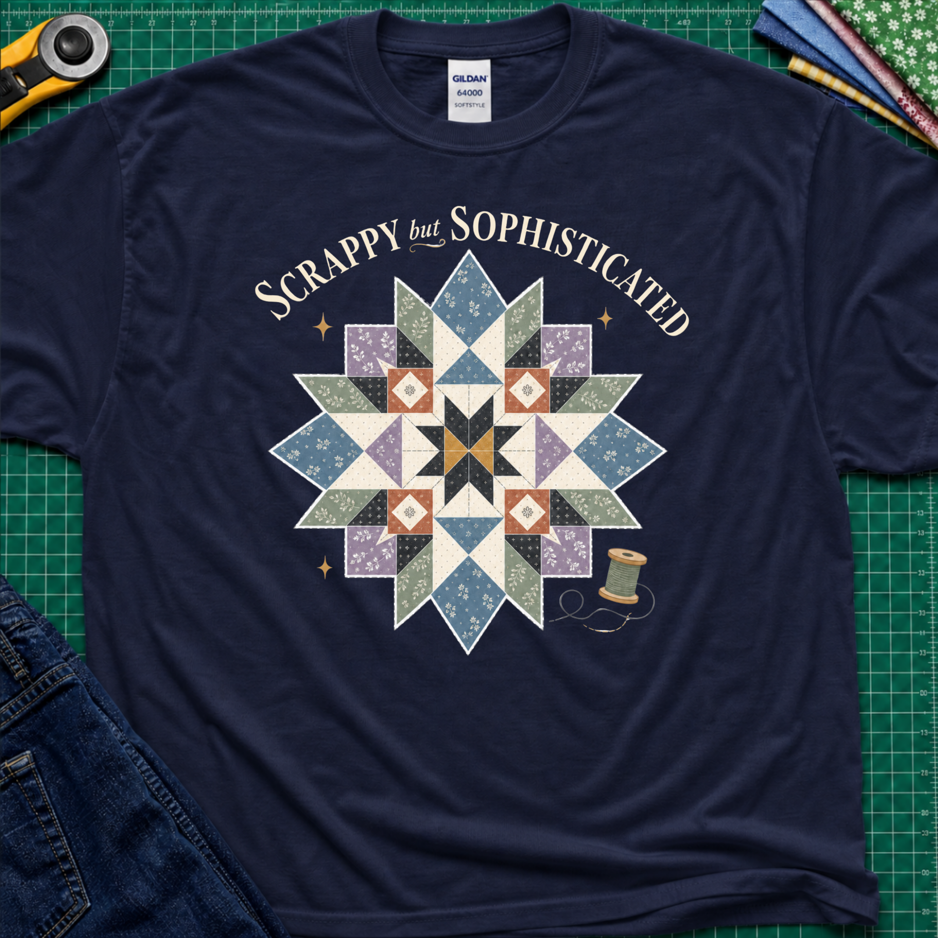 A navy blue Gildan t-shirt with a colorful quilted starburst graphic and the text 'Scrappy but Sophisticated' printed on the front, styled on a green cutting mat with sewing tools and denim.