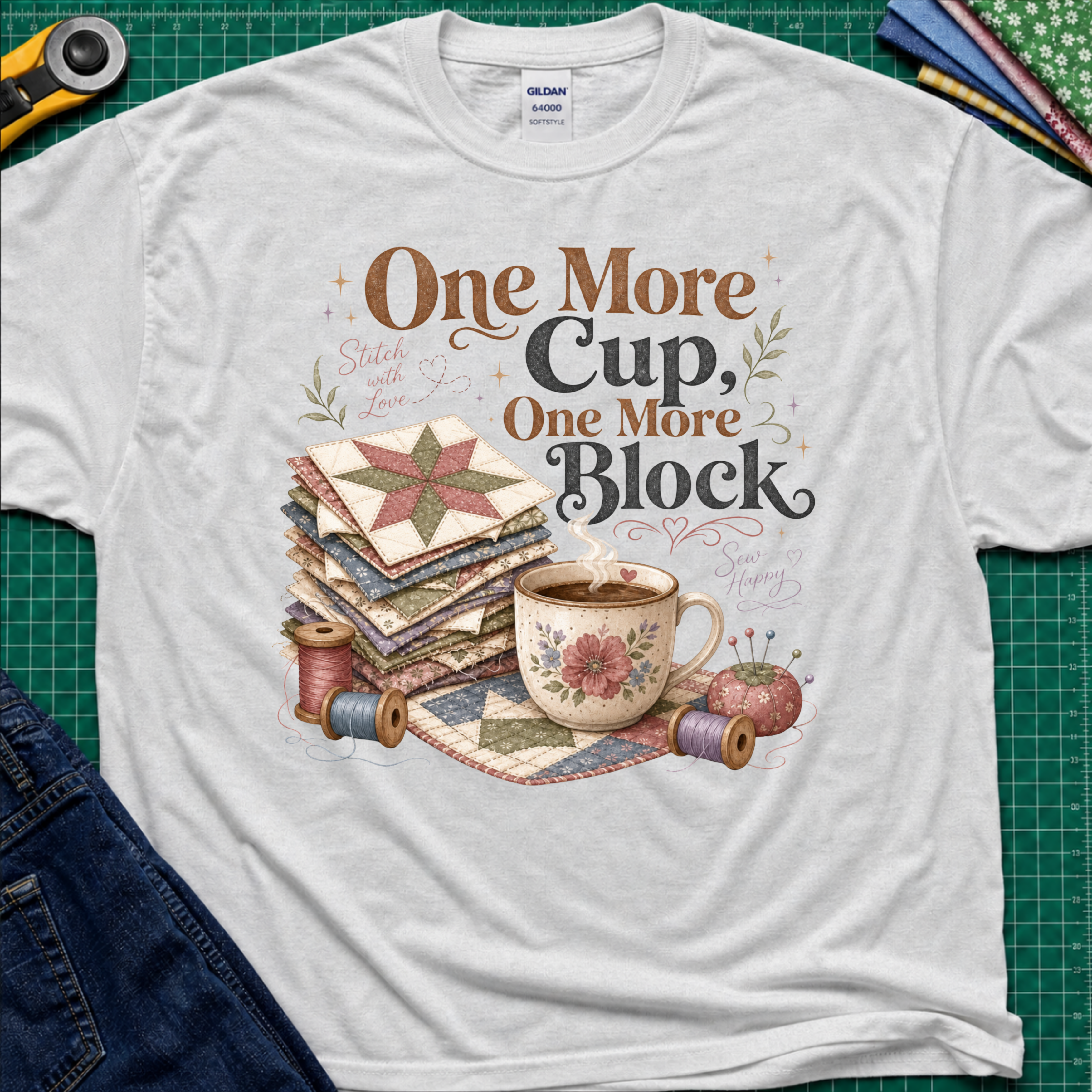 A light grey crewneck t-shirt that reads 'One More Cup, One More Block' above a floral teacup, a stack of quilt blocks, and thread spools. The shirt is laid flat on a green cutting mat with crafting tools partially visible at the edges.