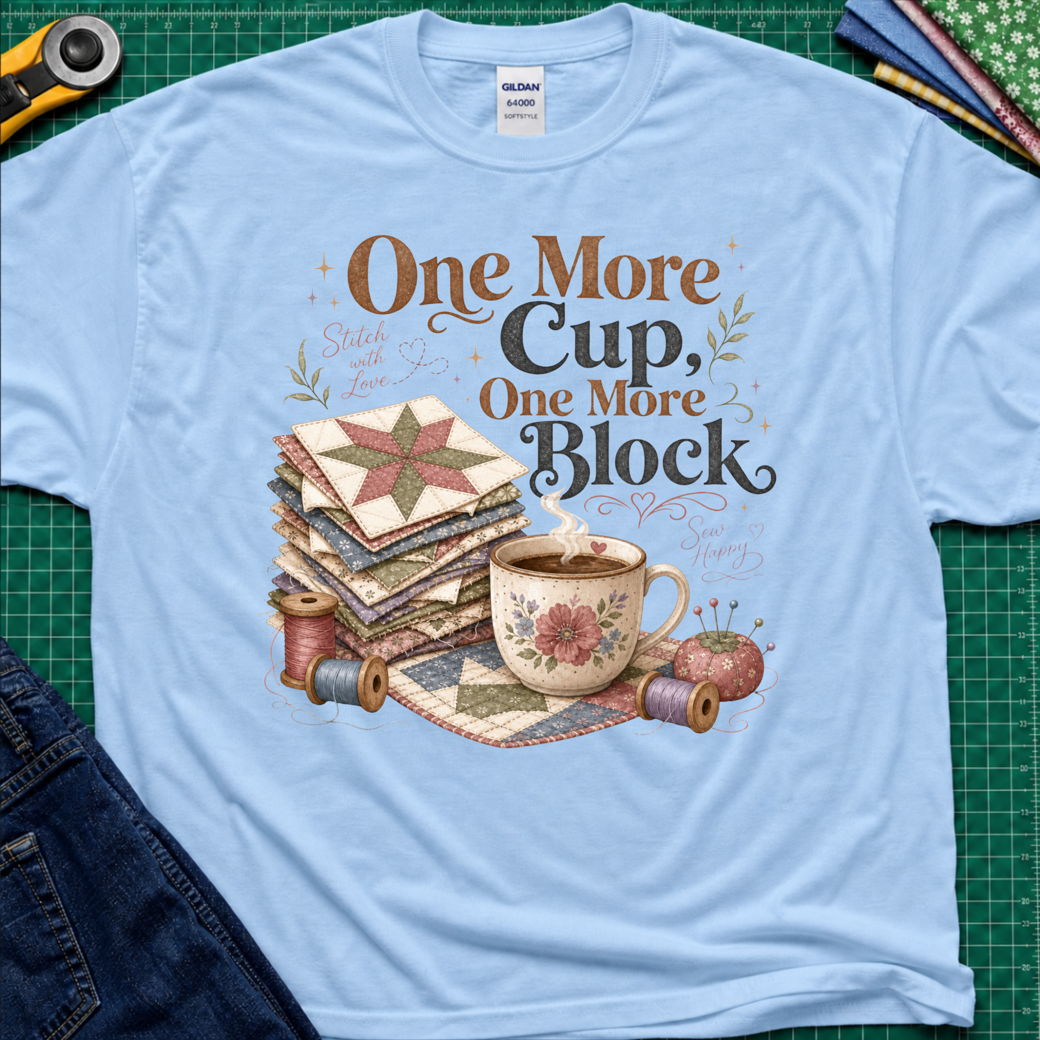 A light blue crewneck t-shirt that reads 'One More Cup, One More Block' above a floral teacup, a stack of quilt blocks, and thread spools. The shirt is laid flat on a green cutting mat with crafting tools partially visible at the edges.