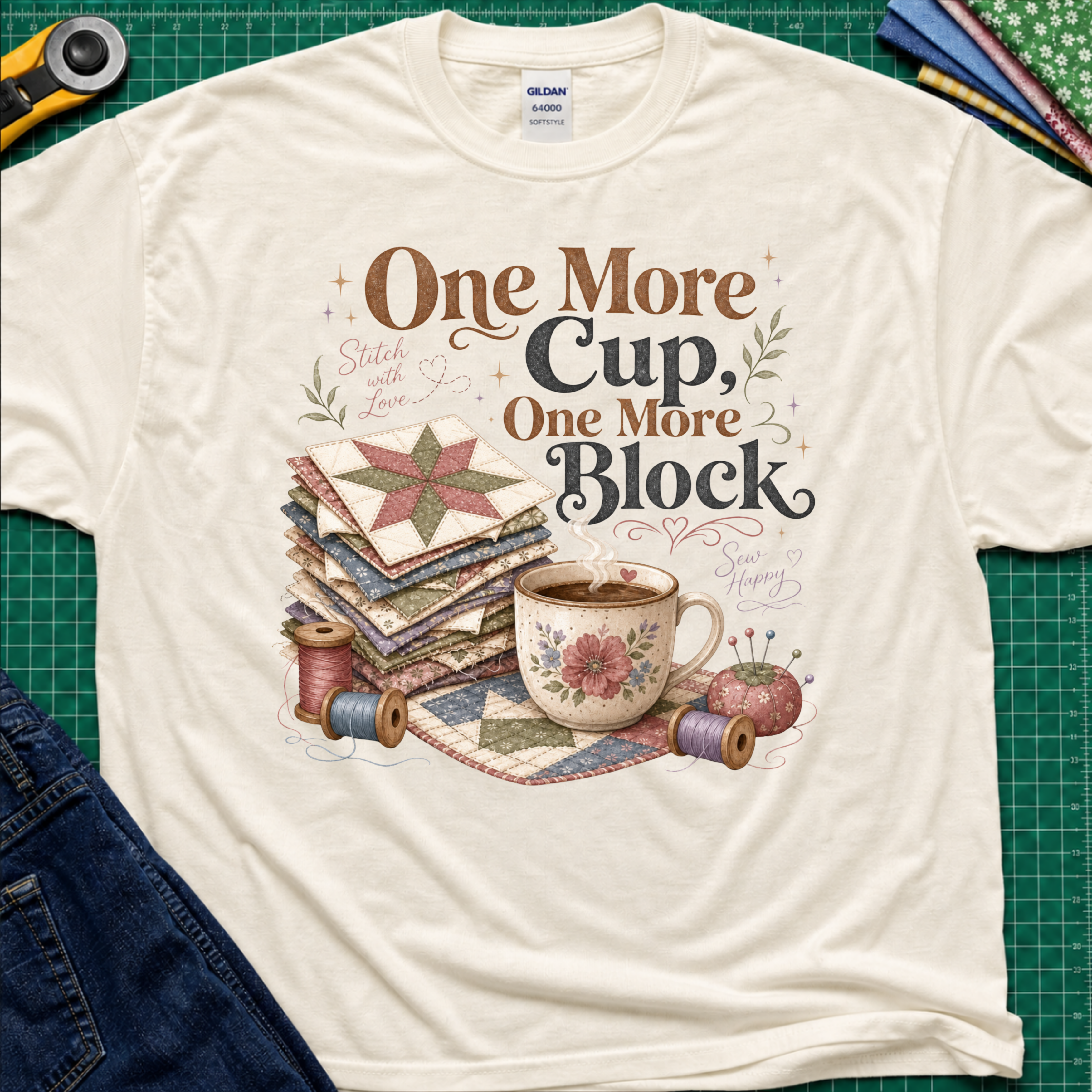 A light cream crewneck t-shirt that reads 'One More Cup, One More Block' above a floral teacup, a stack of quilt blocks, and thread spools. The shirt is laid flat on a green cutting mat with crafting tools partially visible at the edges.