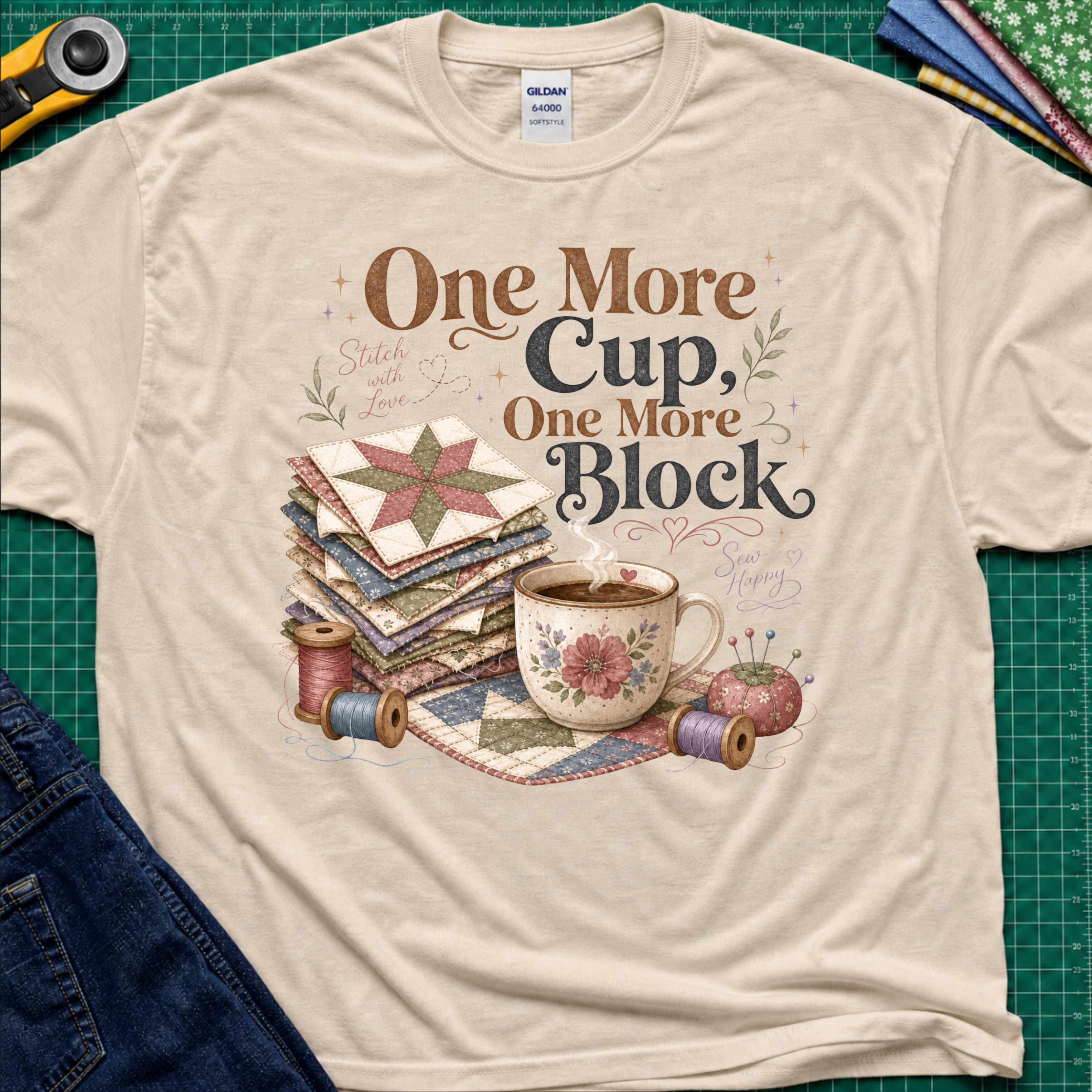 A dark cream crewneck t-shirt that reads 'One More Cup, One More Block' above a floral teacup, a stack of quilt blocks, and thread spools. The shirt is laid flat on a green cutting mat with crafting tools partially visible at the edges.